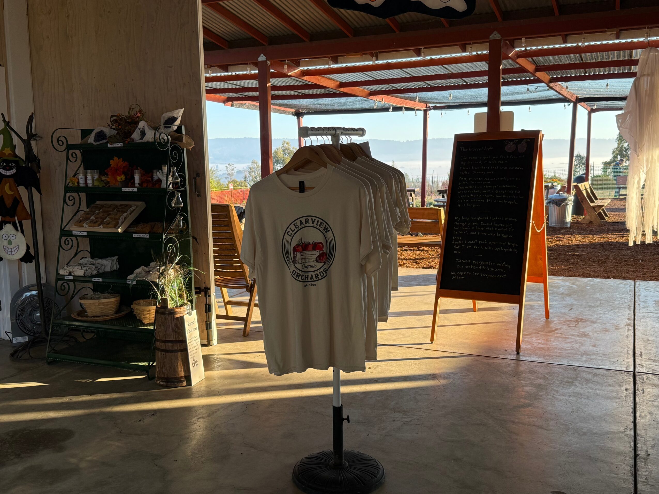 T-shirts hanging on a stand inside a building with a view outside, a chalkboard sign, and outdoor seating area.