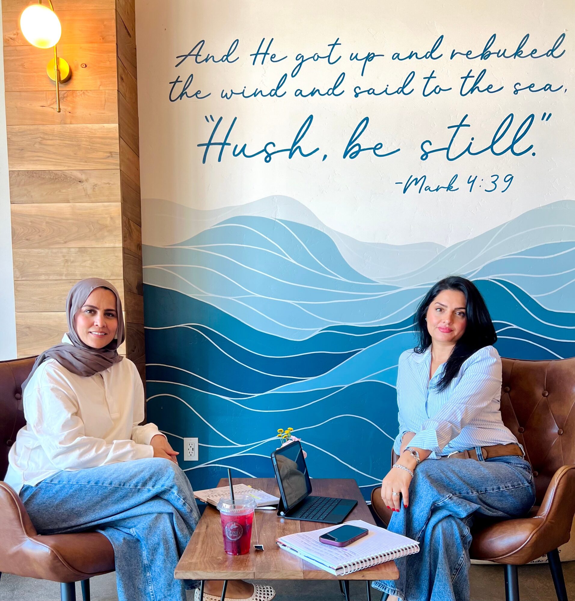 Two women sit on chairs in front of a blue wave mural with a quote from Mark 4:39, holding drinks and notebooks.