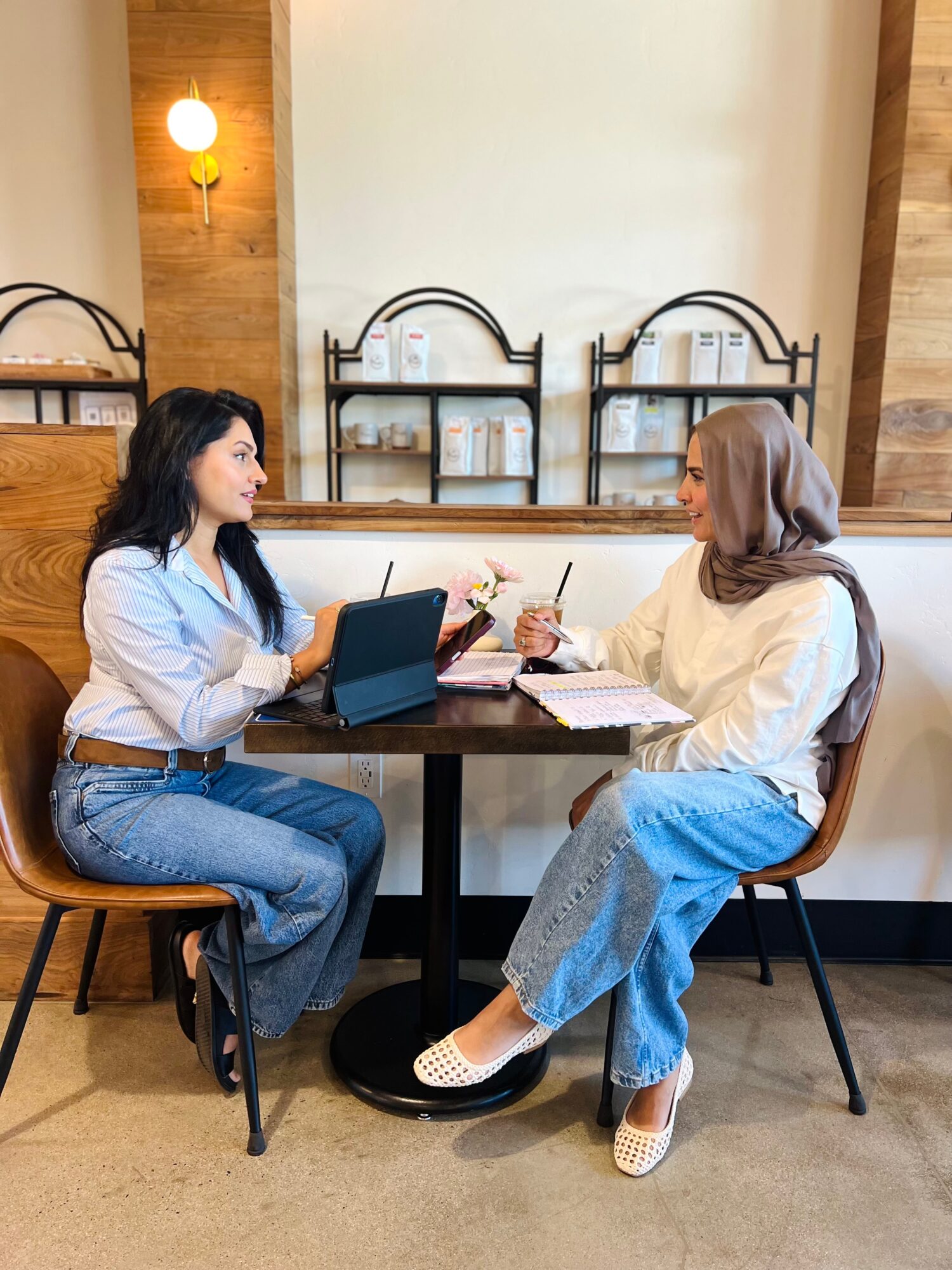 Two women sit at a table in a cafe, talking and using laptops, with wooden wall decor behind them.