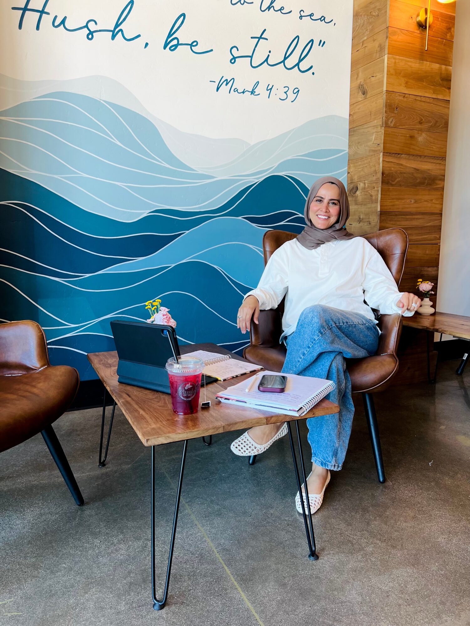 Woman sitting in a chair at a table with a laptop, drink, and phone in a cozy cafe with wooden walls and a mural.