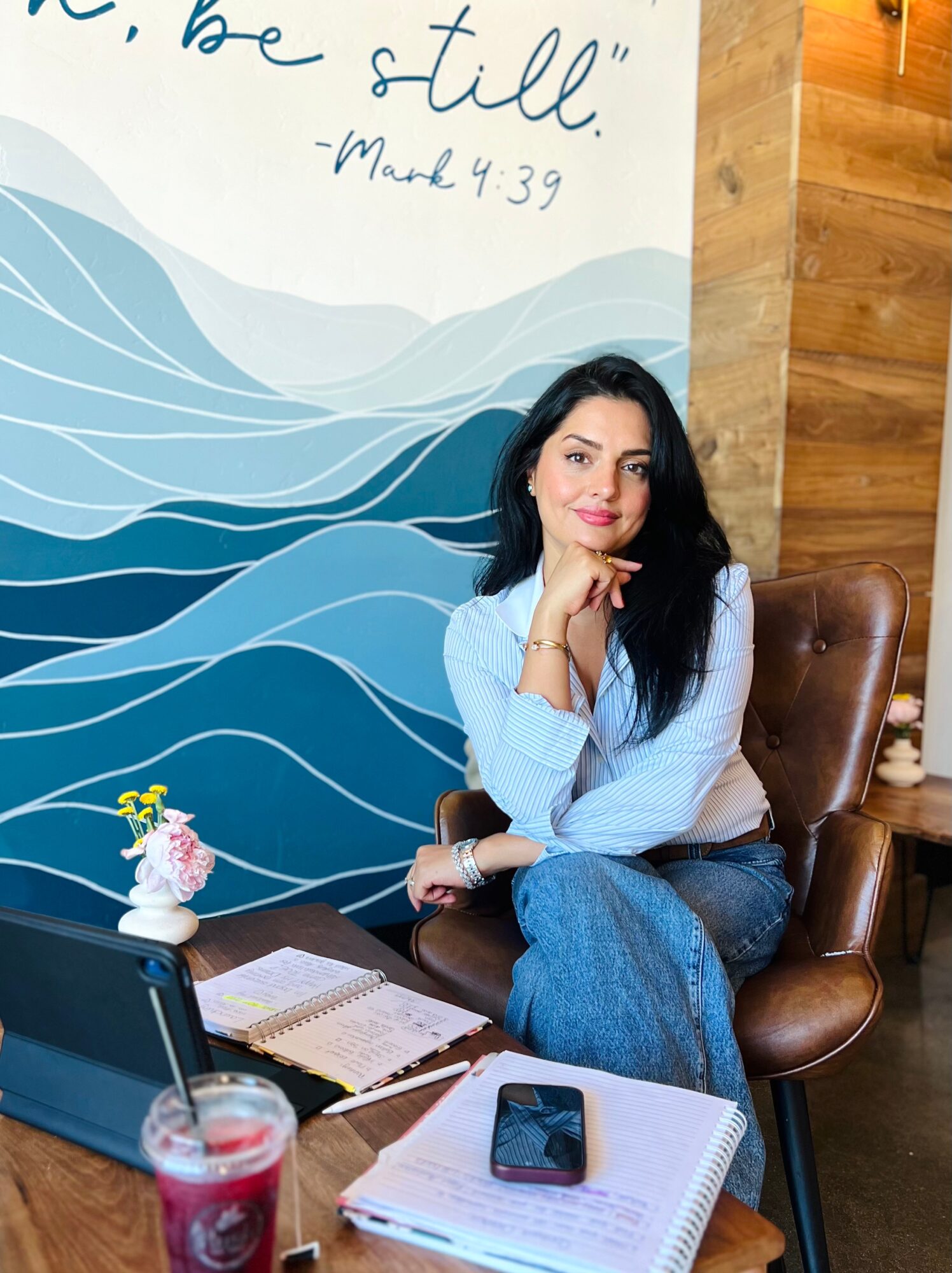 Woman with long dark hair sitting on a brown chair, smiling, in a cafe with a blue wave mural and wooden wall behind her.