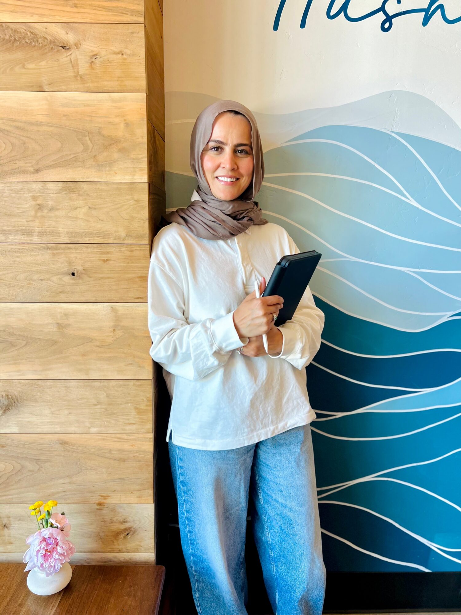 Woman with a headscarf holding a phone, smiling, standing indoors near a wooden wall and a decorative panel.