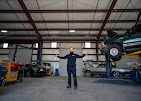 Person standing with arms outstretched inside a large garage or warehouse, with cars and equipment around.