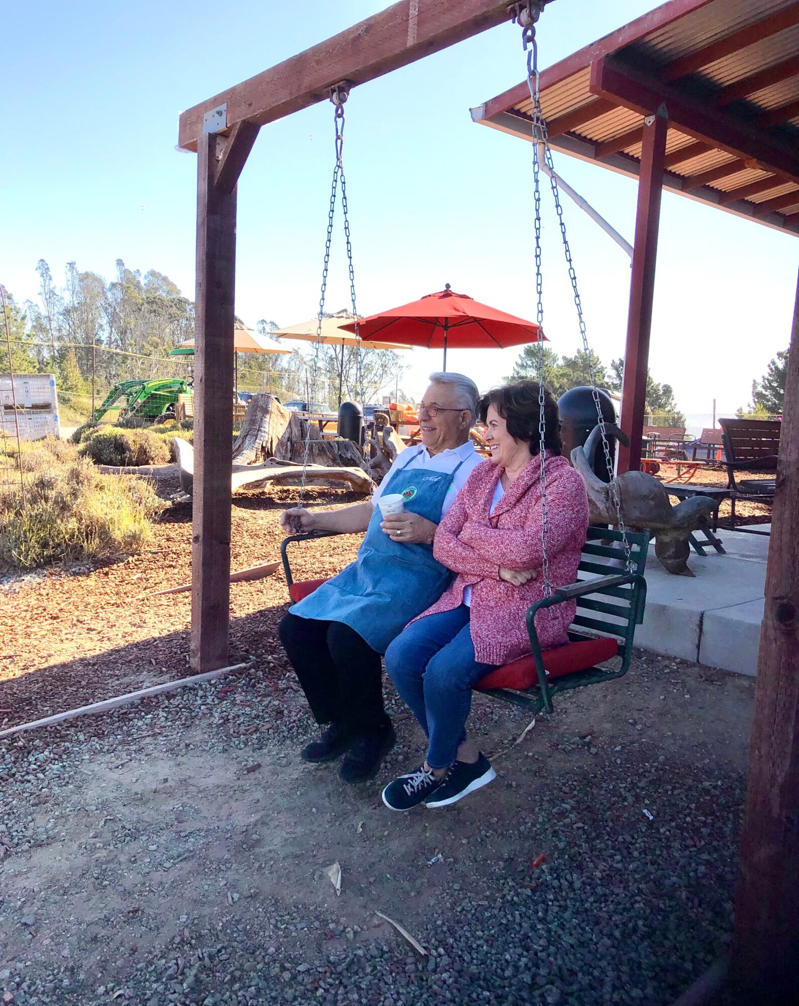 Two people sit on a swing with a red umbrella overhead, outdoors with trees and structures in the background.