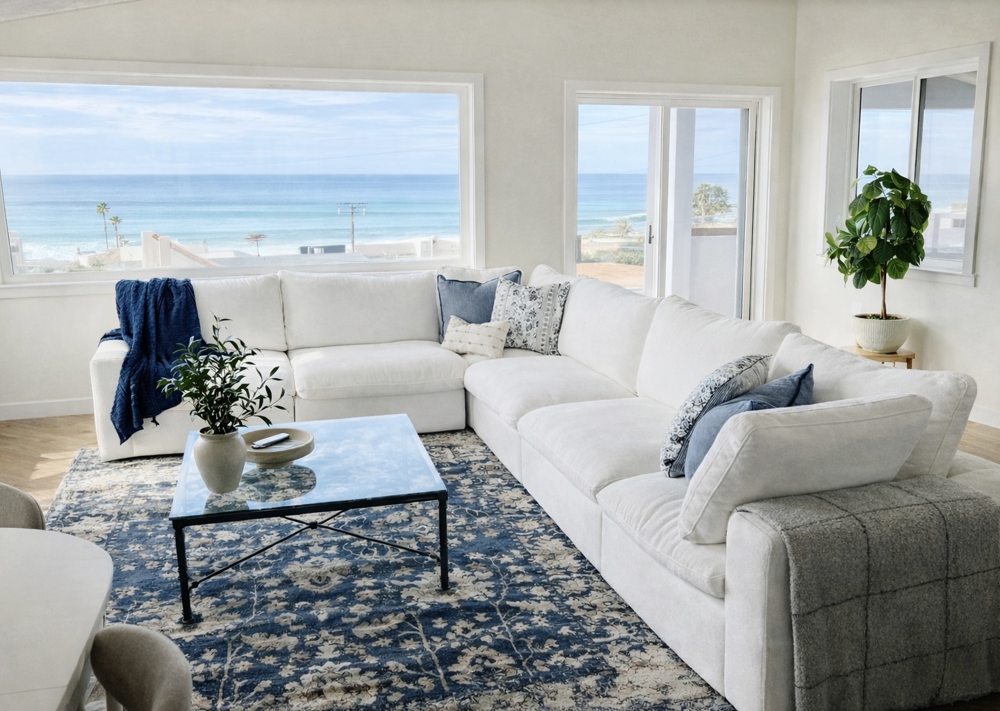 Living room with white sectional sofa, glass coffee table, blue and white pillows, large windows showing beach view, potted plant.