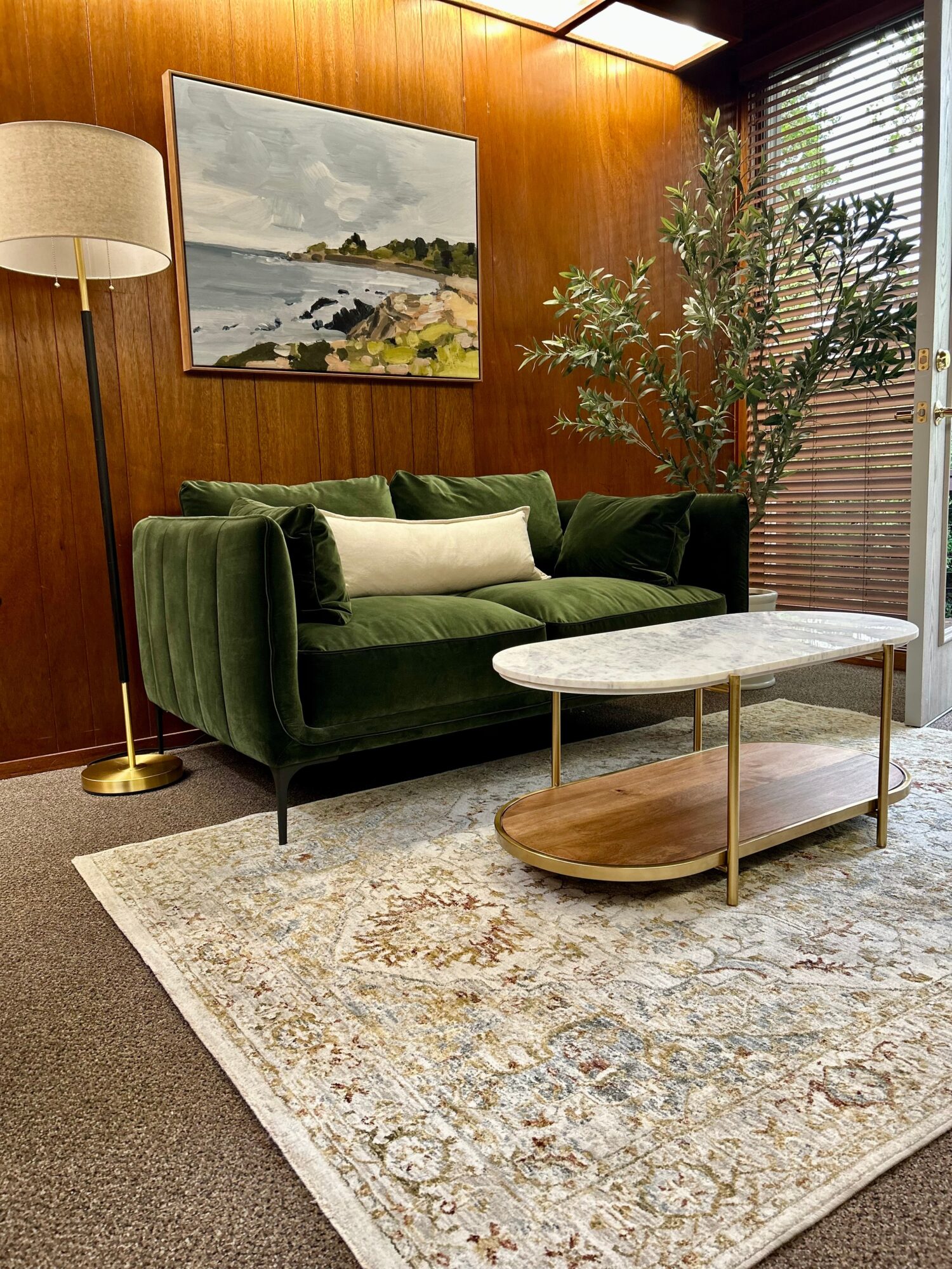 Living room with green velvet sofa, marble coffee table, large potted plant, painting, and window with blinds.