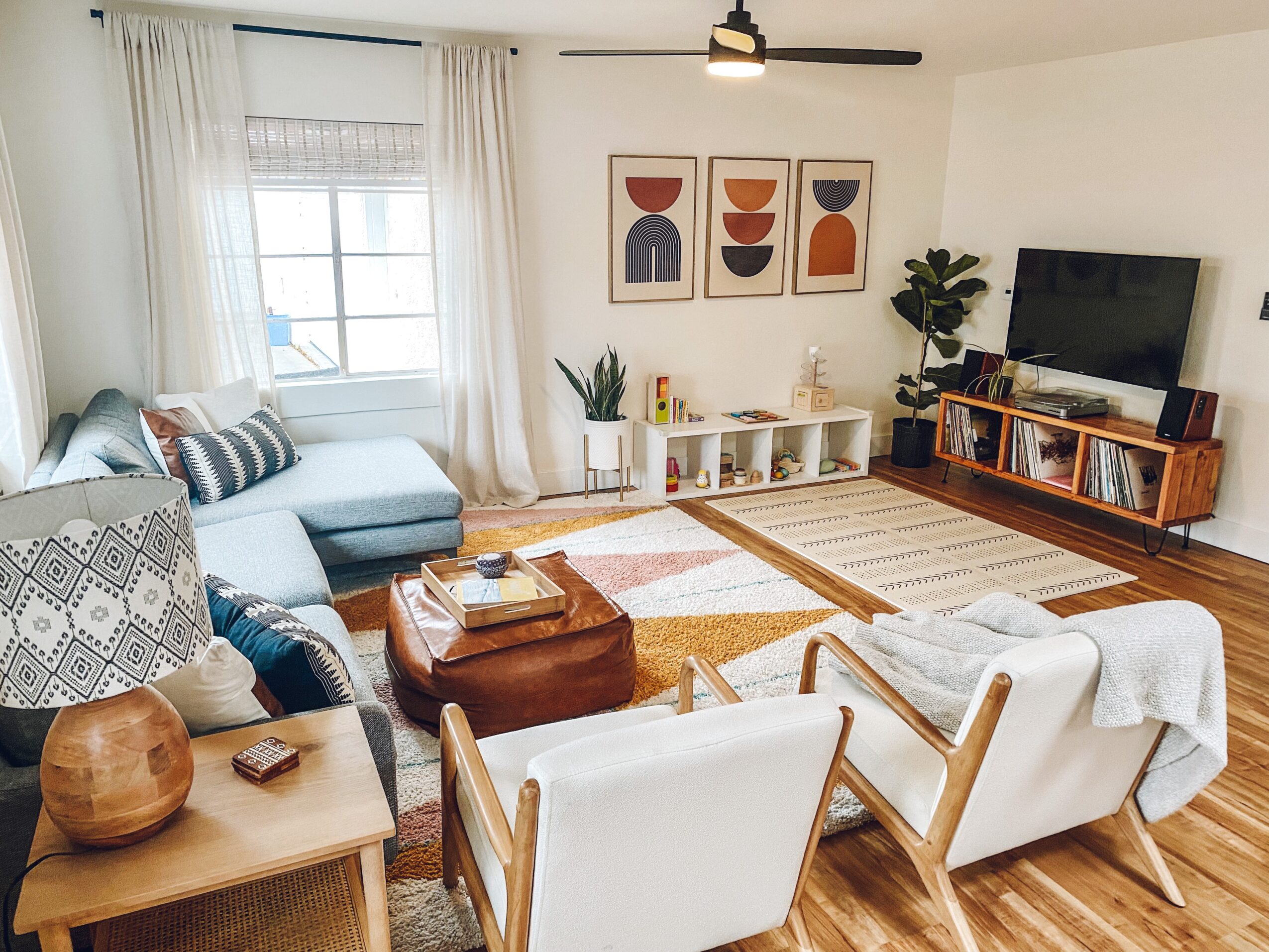 Living room with white walls, large window, sofa, armchairs, TV, bookshelf, and artwork, decorated with plants and rugs.
