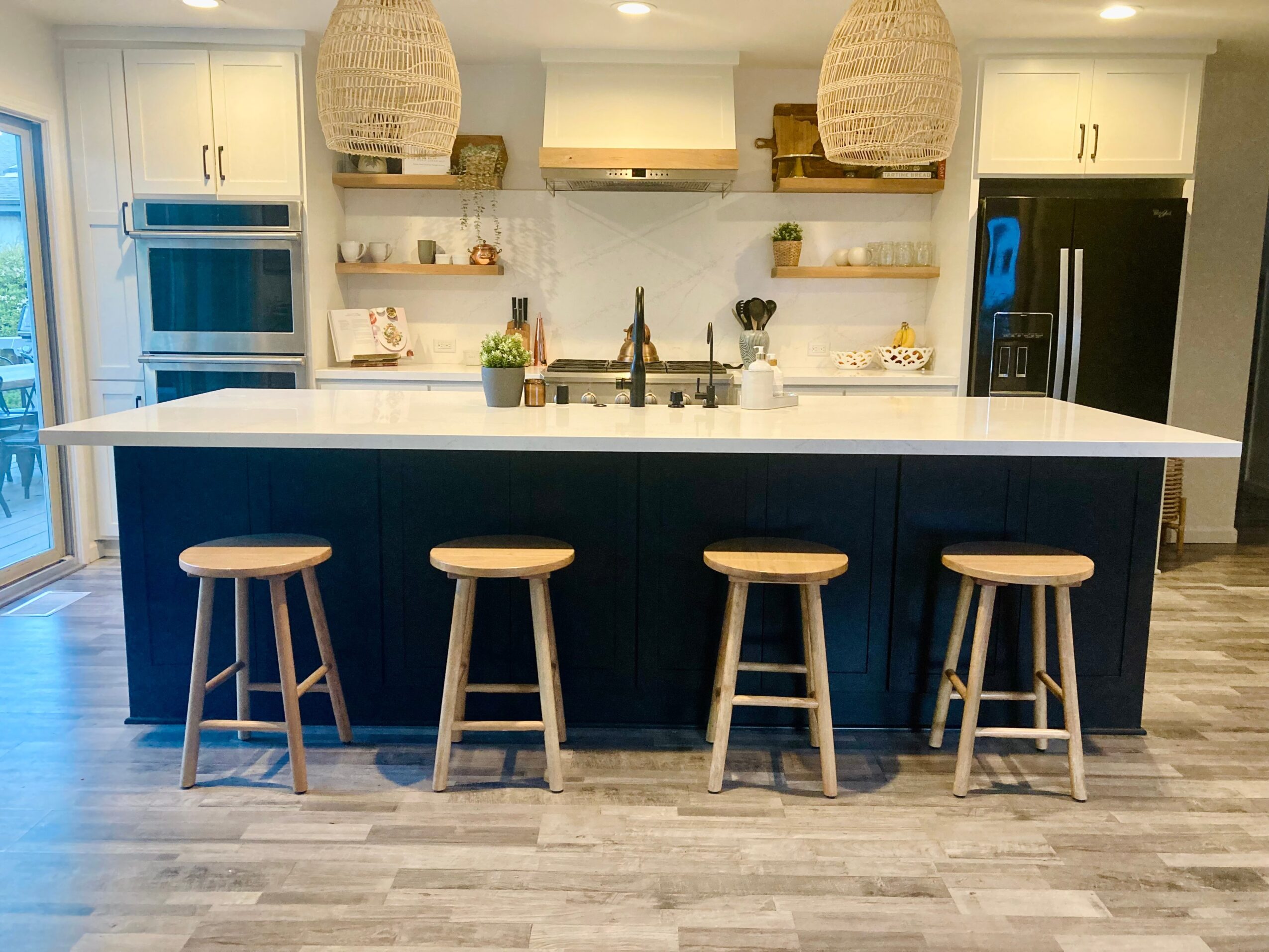 Modern kitchen with a large island, four wooden stools, white cabinets, open shelves, and pendant lights.