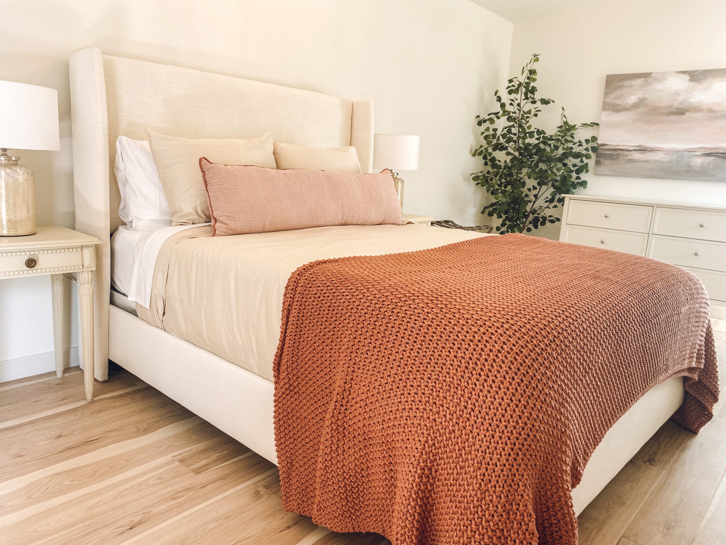 Upright bedroom with a bed, beige headboard, pink and beige pillows, brown blanket, white nightstands, and a dresser with a plant and artwork.