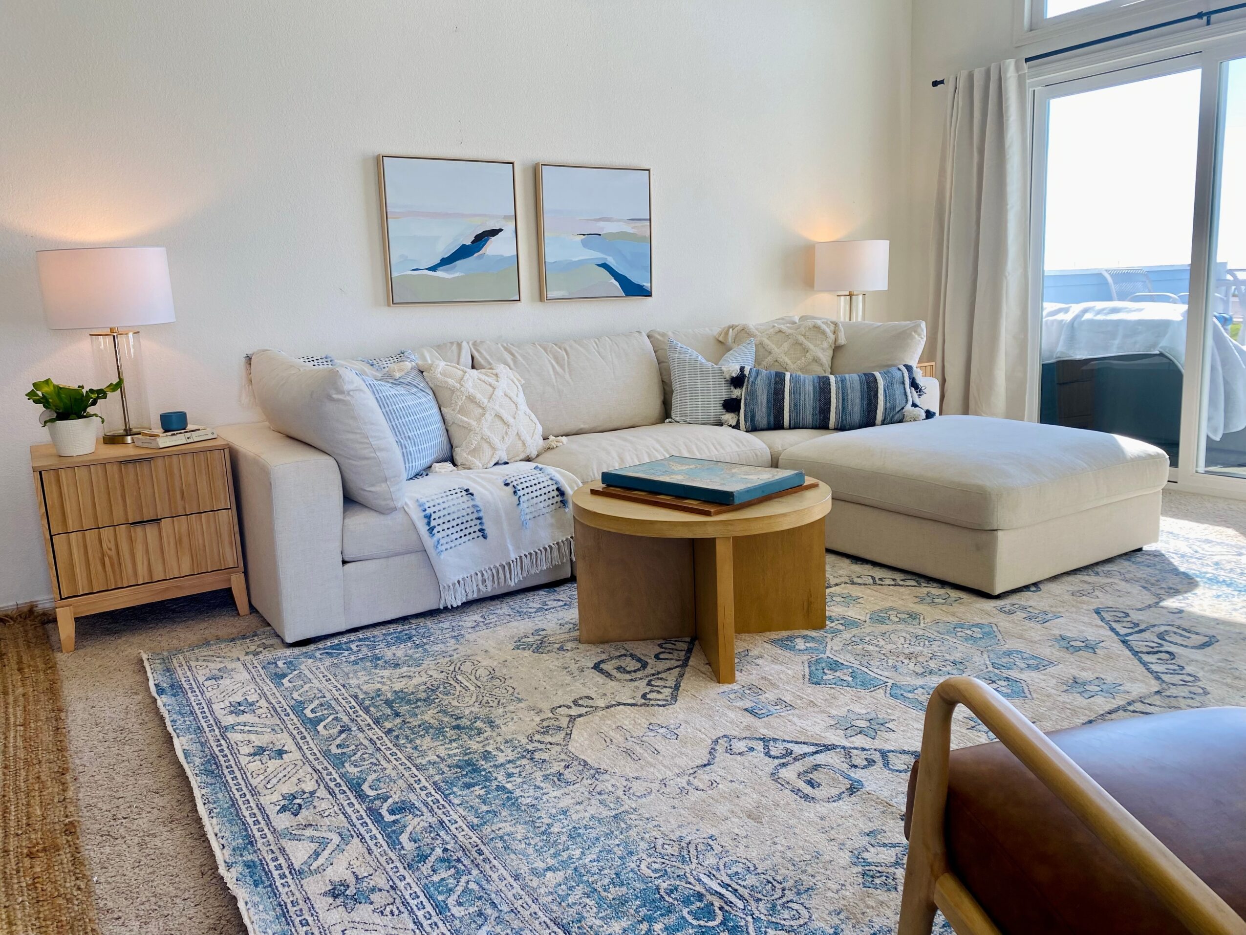 Living room with a white sofa, blue and white pillows, wooden side table, round coffee table, and large window with curtains.