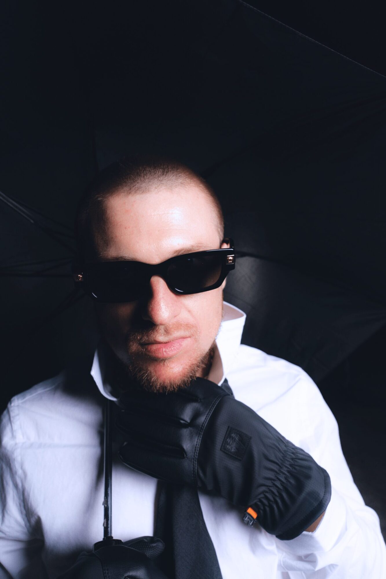 Person wearing sunglasses, white shirt, black gloves, and a black tie, adjusting tie with hand, dark background.