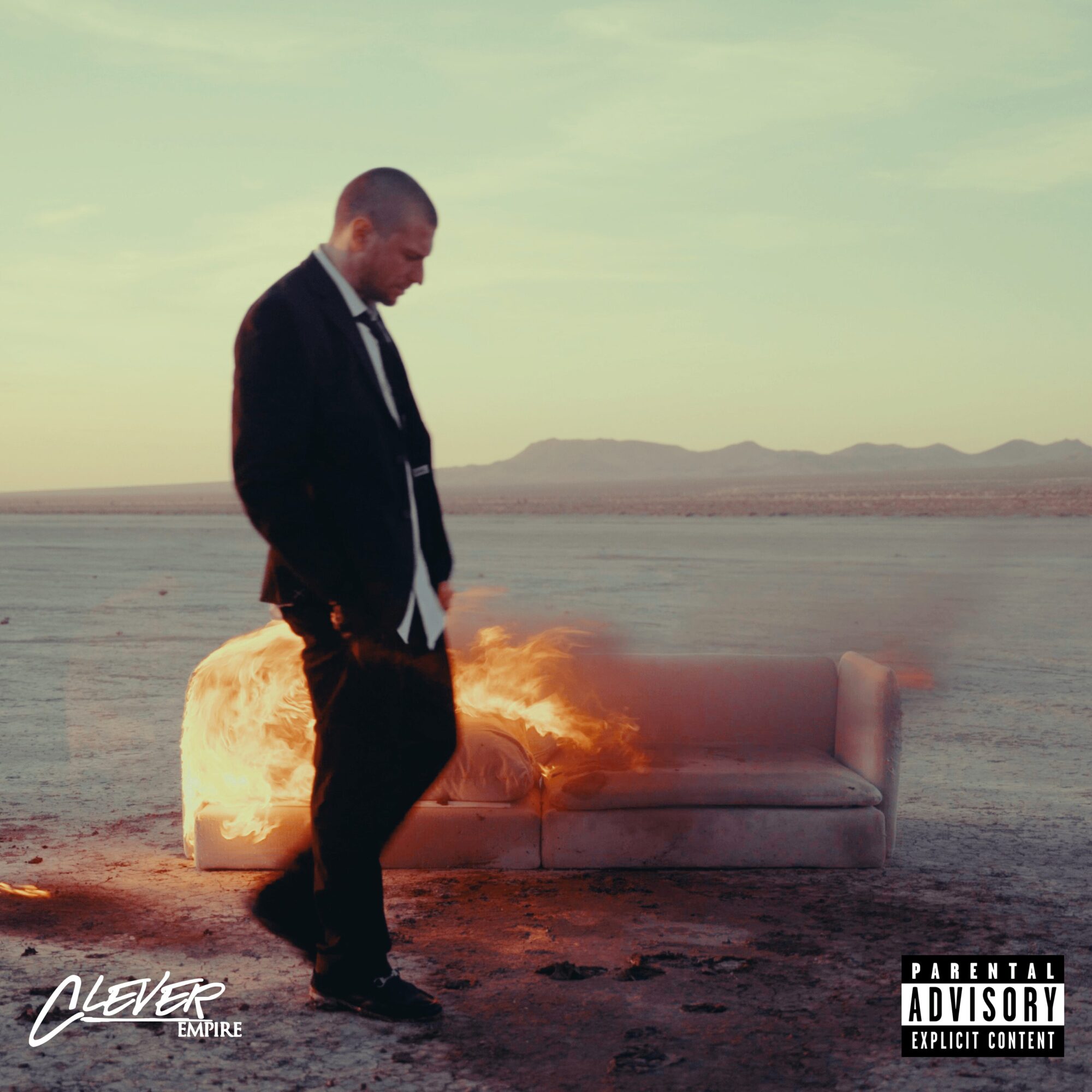 Man in suit walking away from a burning sofa in a desert landscape during sunset.