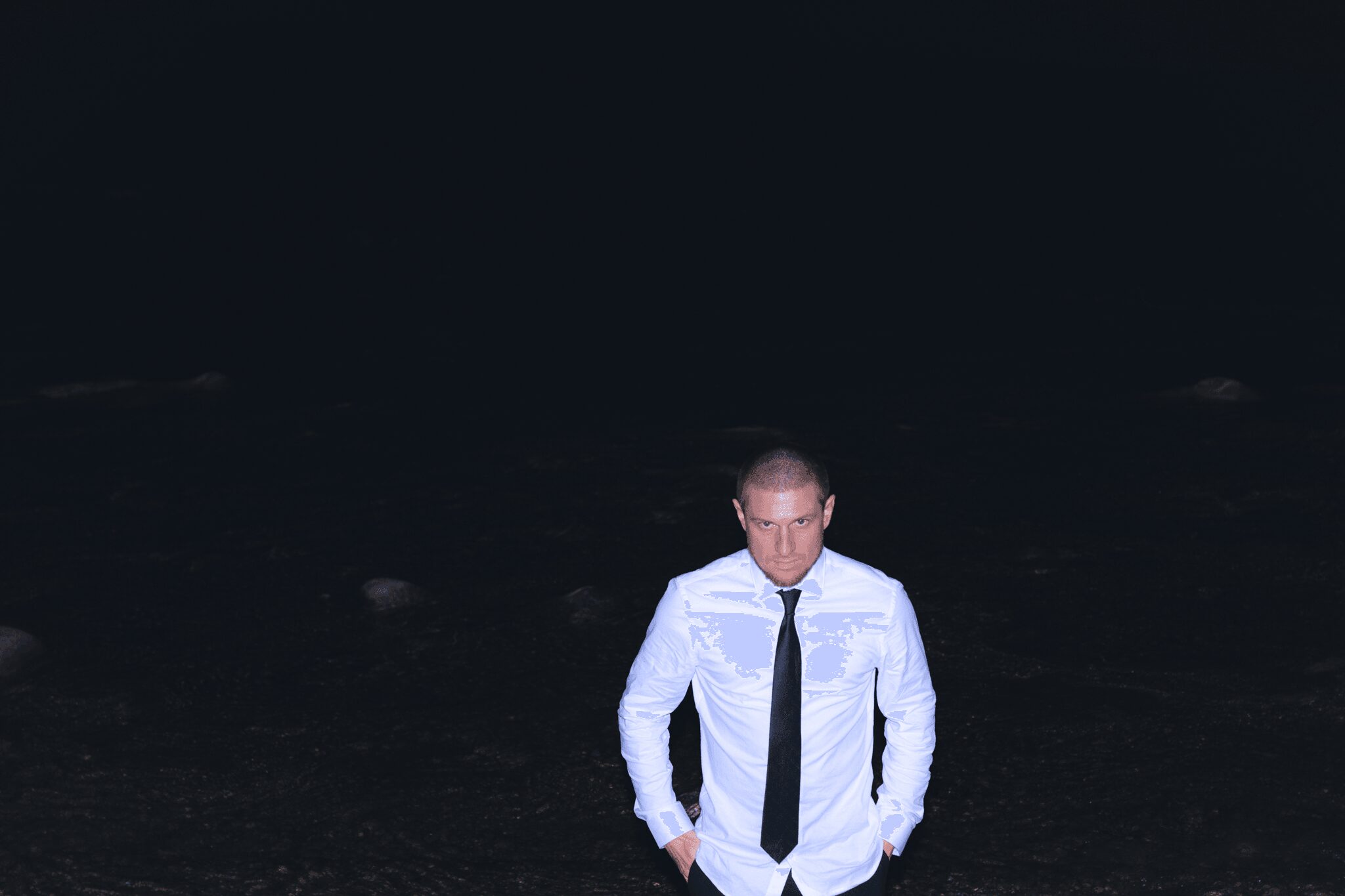 Man in white shirt and black tie standing outdoors at night, hands in pockets, dark background.