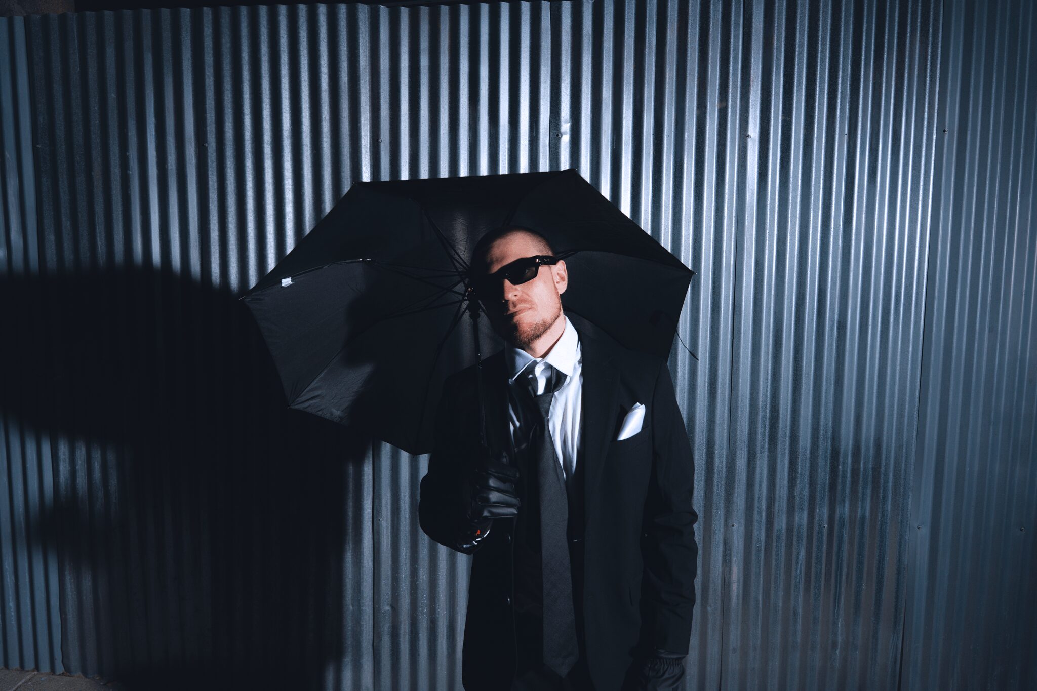 Man in a suit holding an umbrella, standing against a corrugated metal wall, wearing sunglasses, with a serious expression.