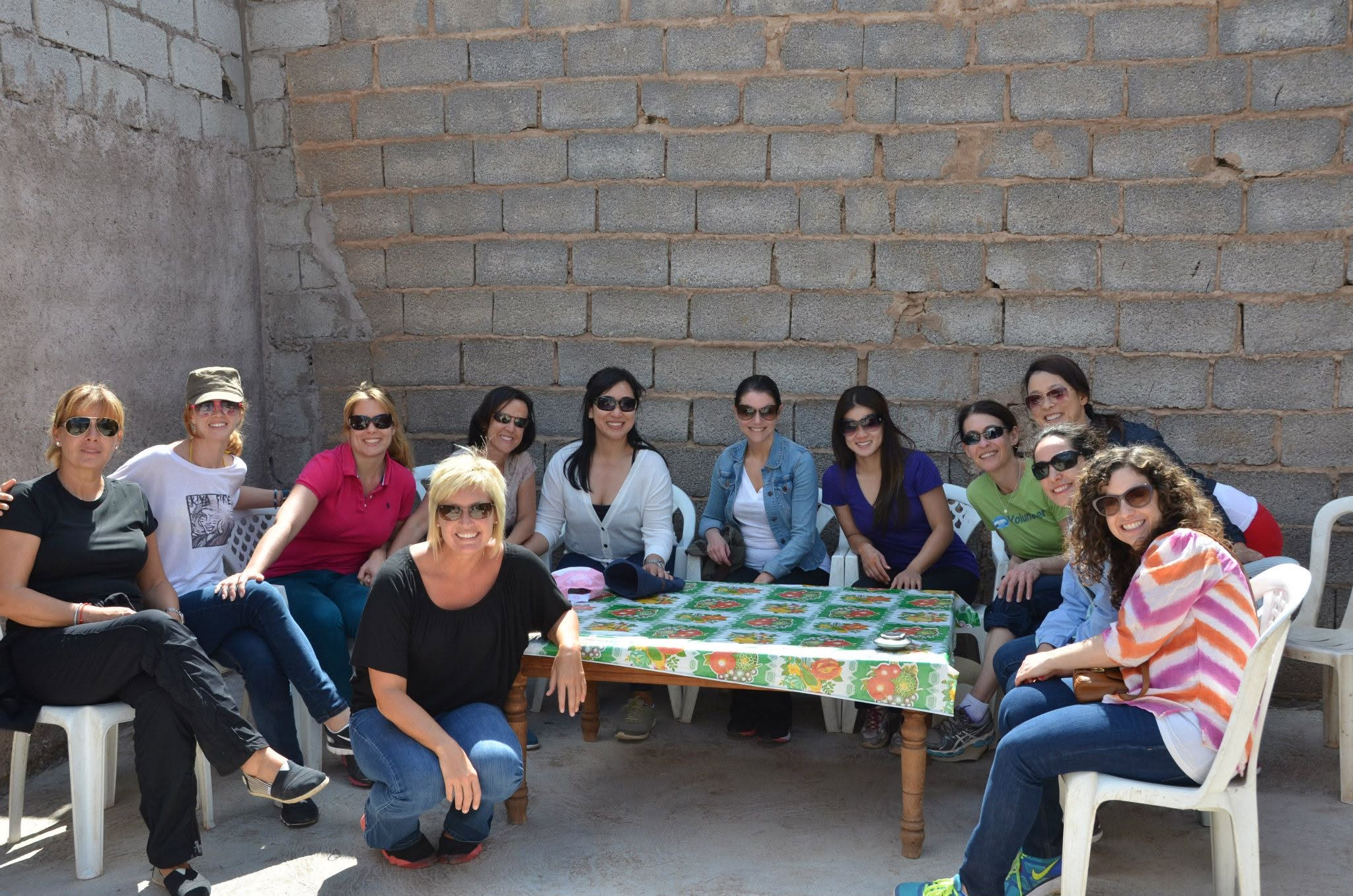 Group of ten women sitting outdoors around a table with a colorful tablecloth, against a brick wall.