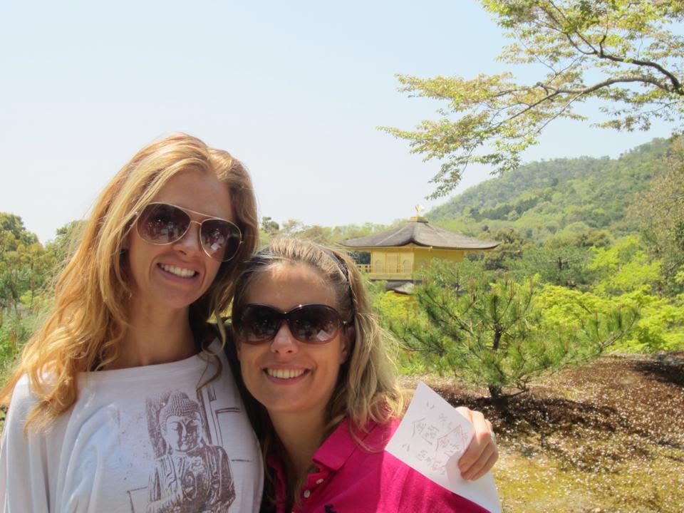 Two women smiling outdoors, wearing sunglasses, with a scenic background of trees and a building, one in a white shirt and the other in pink.