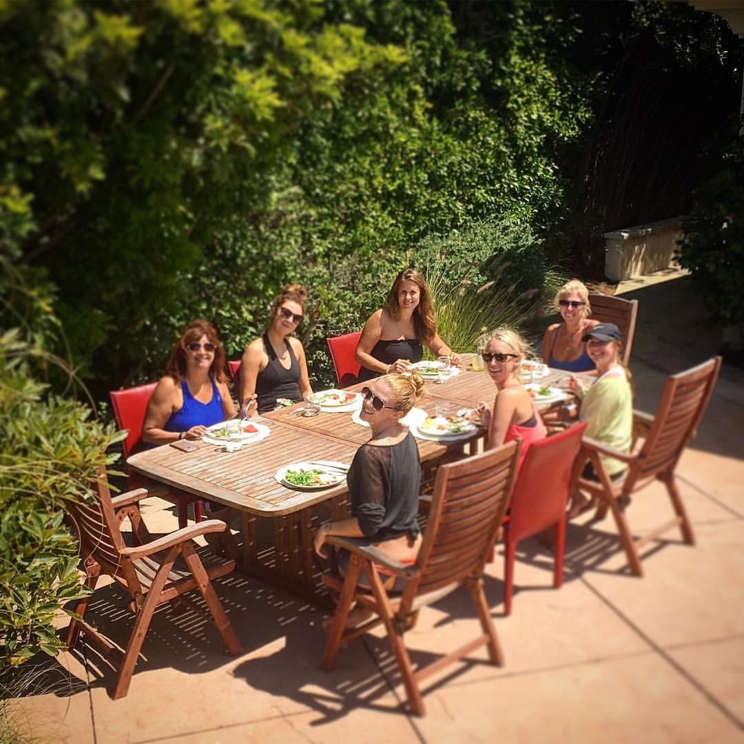 Group of seven people sitting around a wooden outdoor table with food, surrounded by greenery.