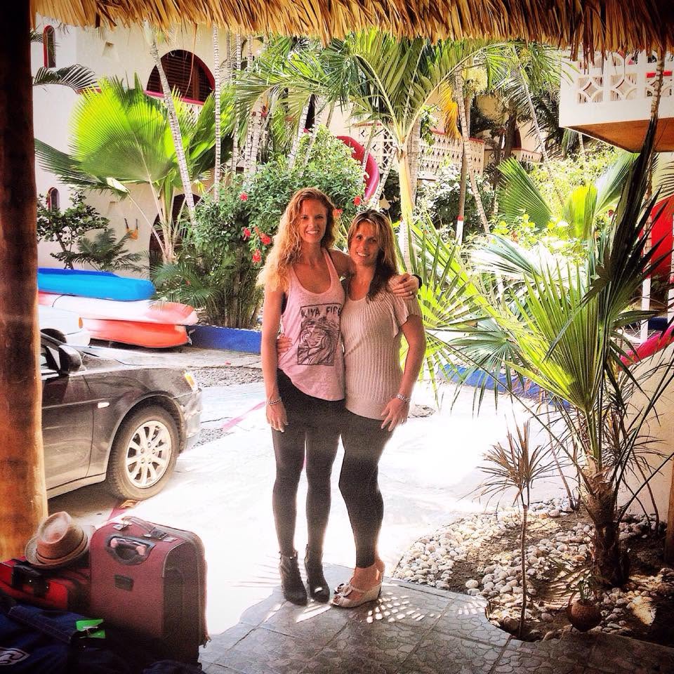 Two women stand together outdoors with tropical plants and boats in the background, smiling at the camera.