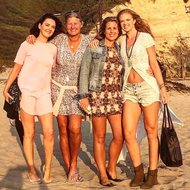 Four women standing together outdoors, smiling, with a sandy background and trees, dressed in casual summer clothing.