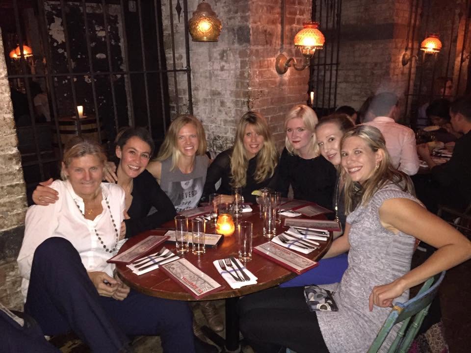 Group of seven women sitting around a table in a dimly lit restaurant, smiling and posing for the photo.