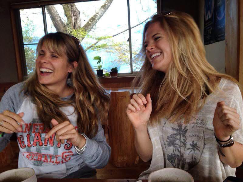 Two women laughing and smiling indoors near a window with trees outside.