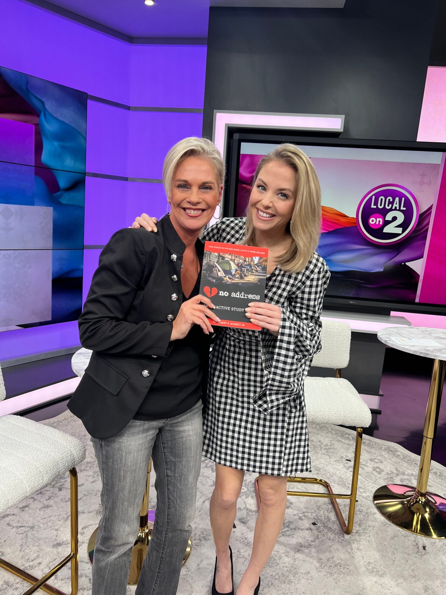 Two women sitting on a white couch in a TV studio, one holding a book, with a TV screen in the background.