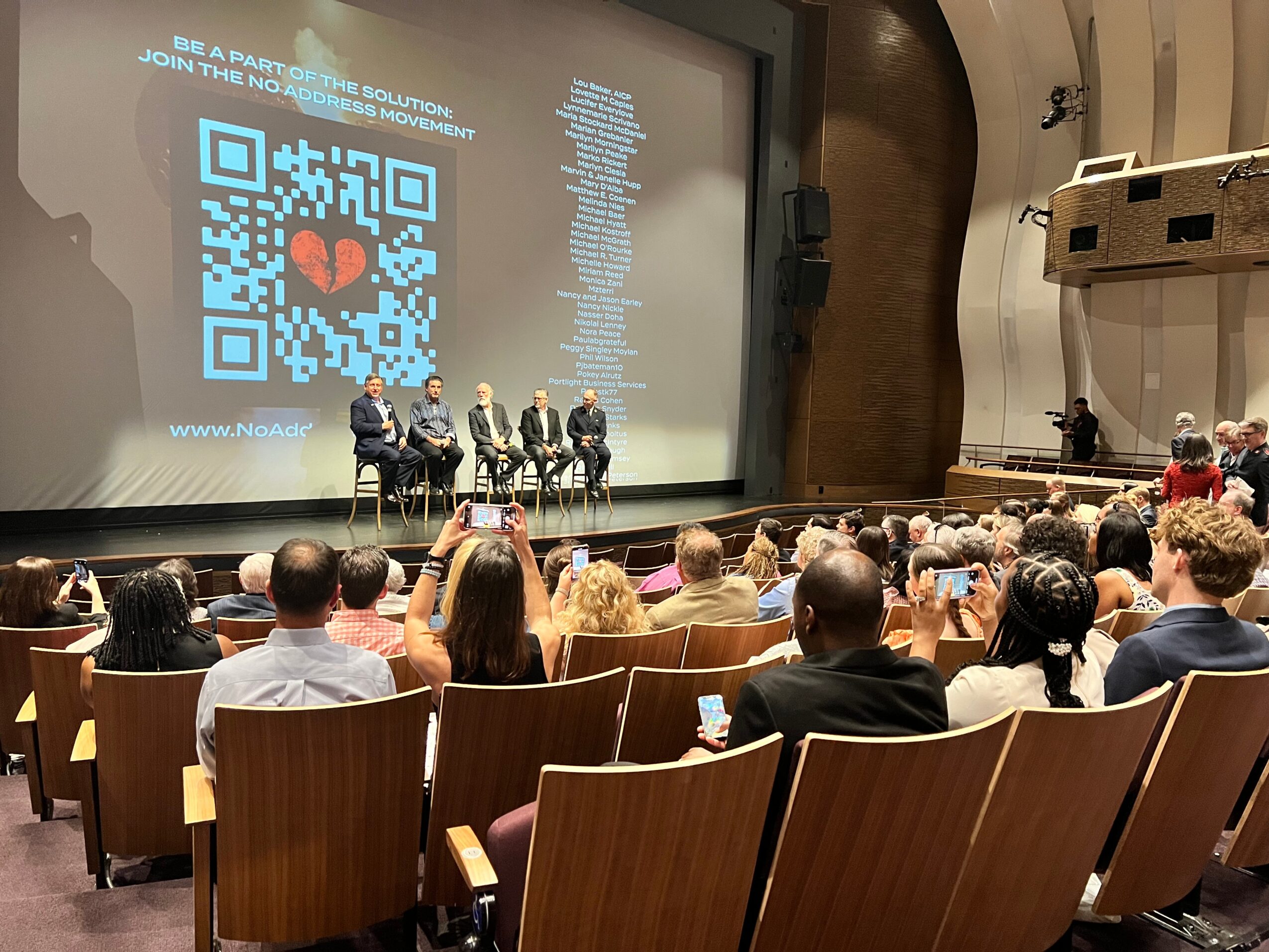Panel discussion on stage with four people, audience seated in a theater, large screen with QR code and text behind them.