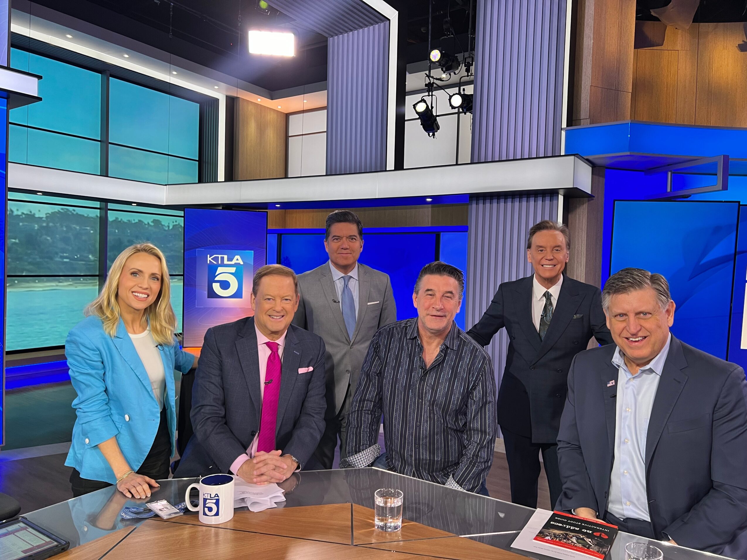 Six news anchors and hosts in a studio, smiling, with a cityscape background and a KTLA 5 logo, behind a news desk.
