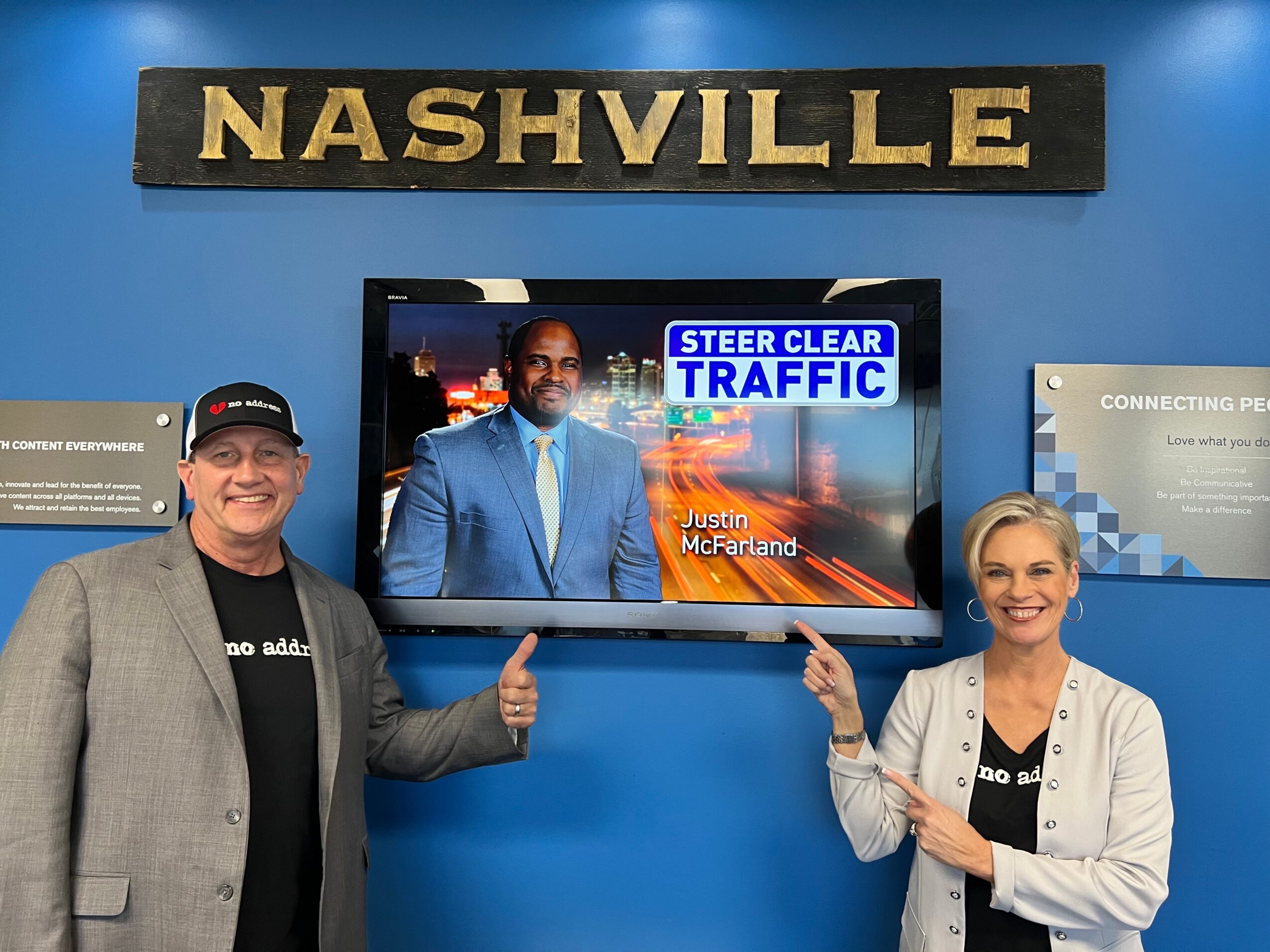 Two people standing in front of a screen with traffic news, smiling and pointing at it, with a Nashville sign above.