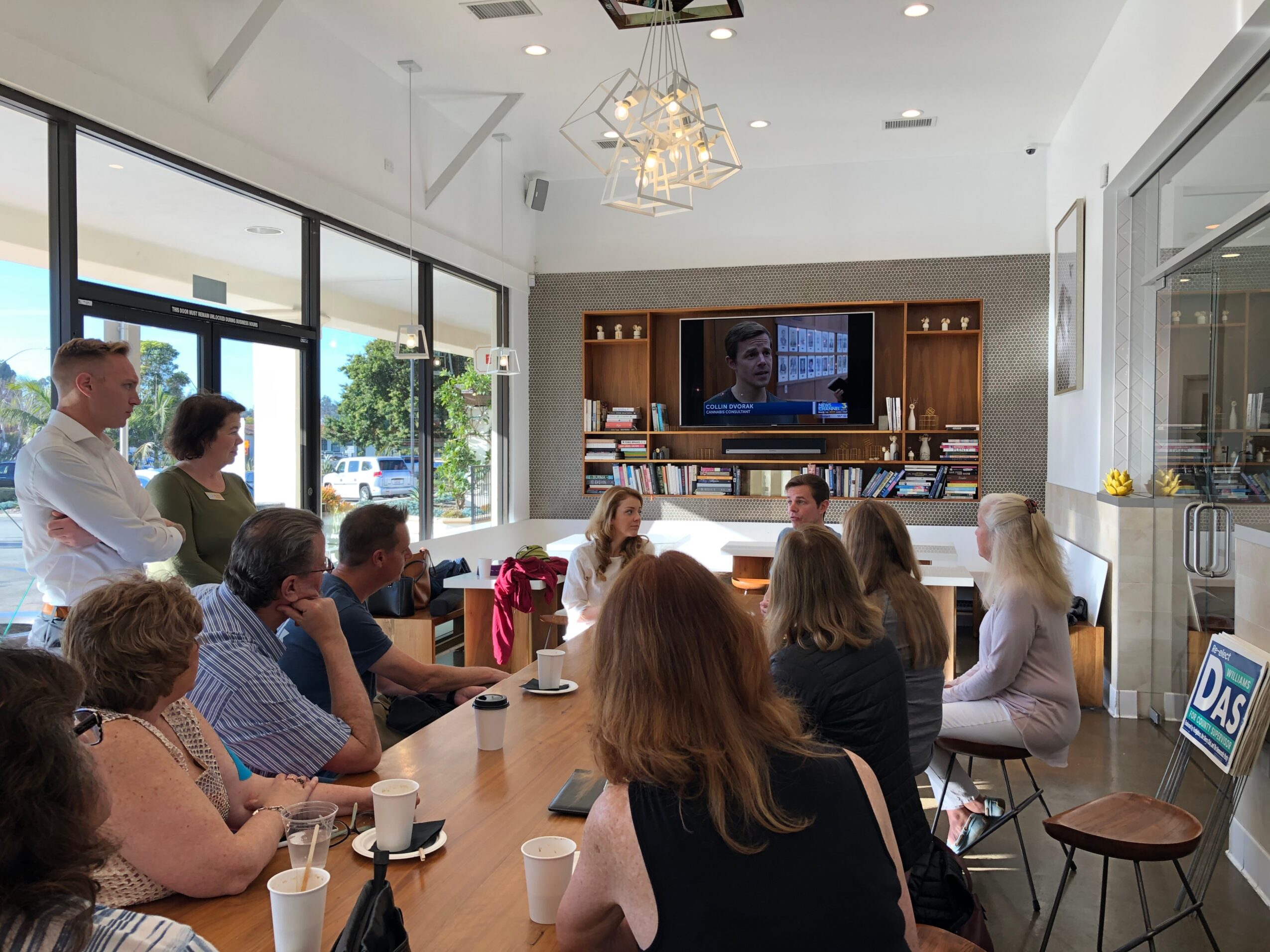 Group of people seated around a table in a bright room watching a television screen, with large windows and modern decor.