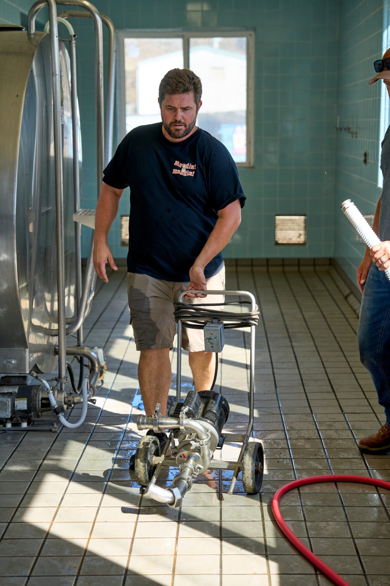 Man walking with a walker in a tiled room, possibly a hospital or clinic, with a person holding a clipboard nearby.
