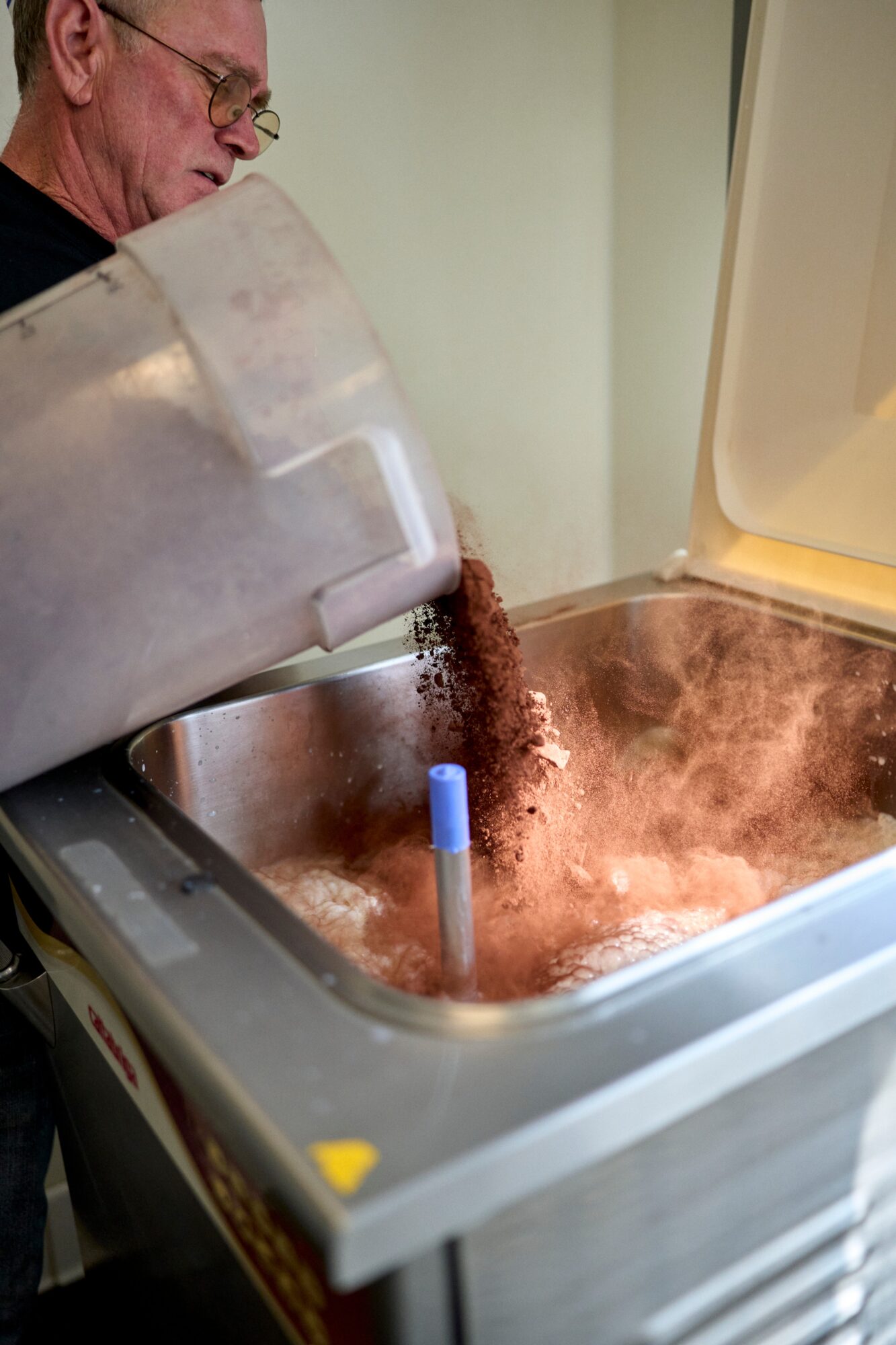 Person pouring coffee beans into a coffee machine, with steam rising from the machine's open lid.