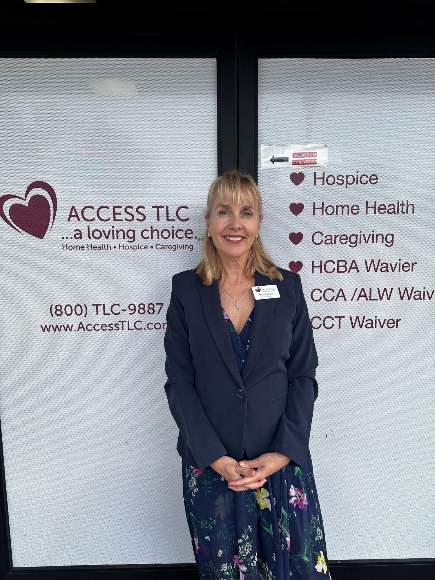 Woman standing in front of a glass door with text about healthcare services, smiling, wearing a dark blazer and floral skirt.