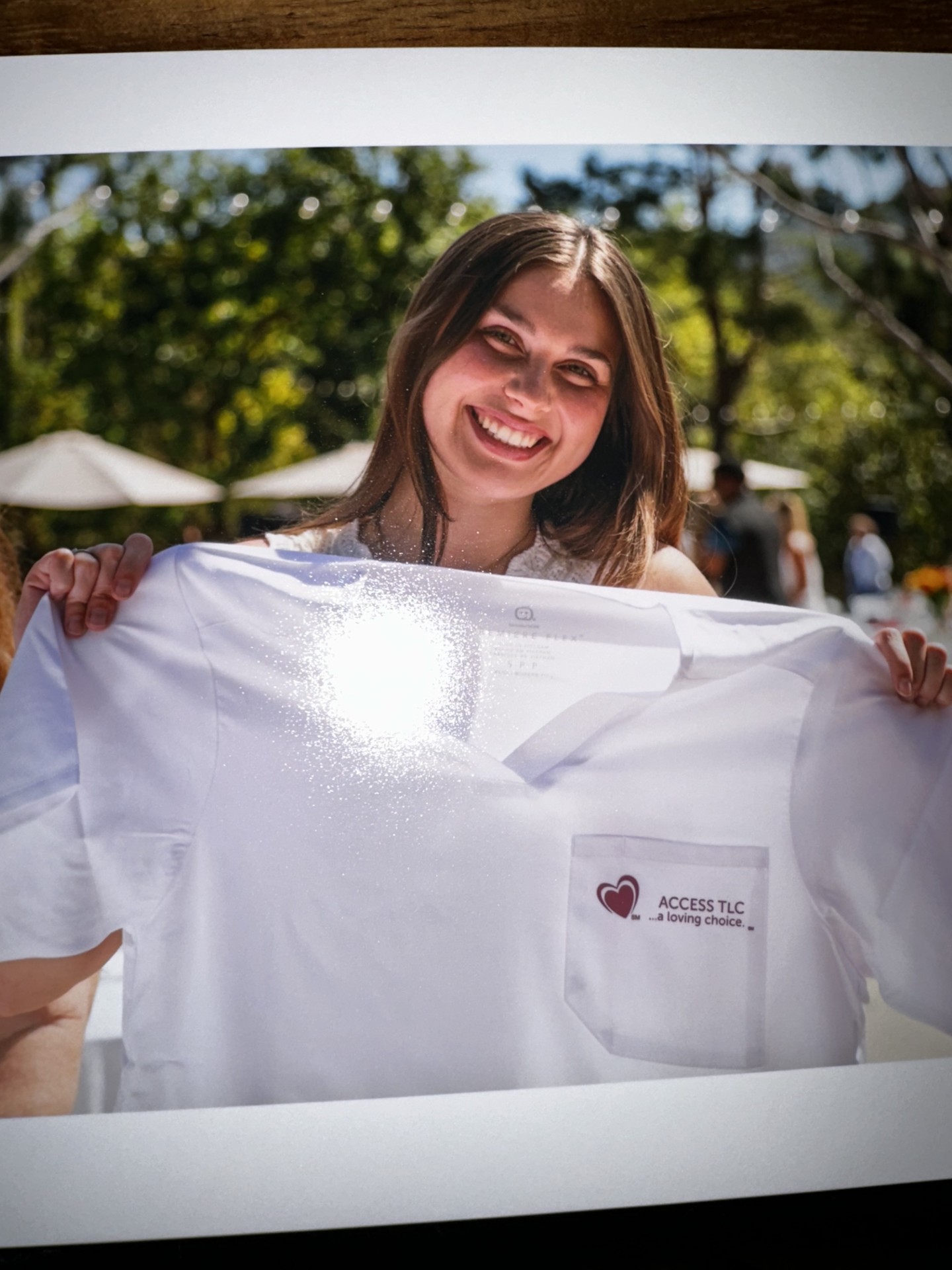 Smiling woman holding a white shirt outdoors with trees and umbrellas in background.