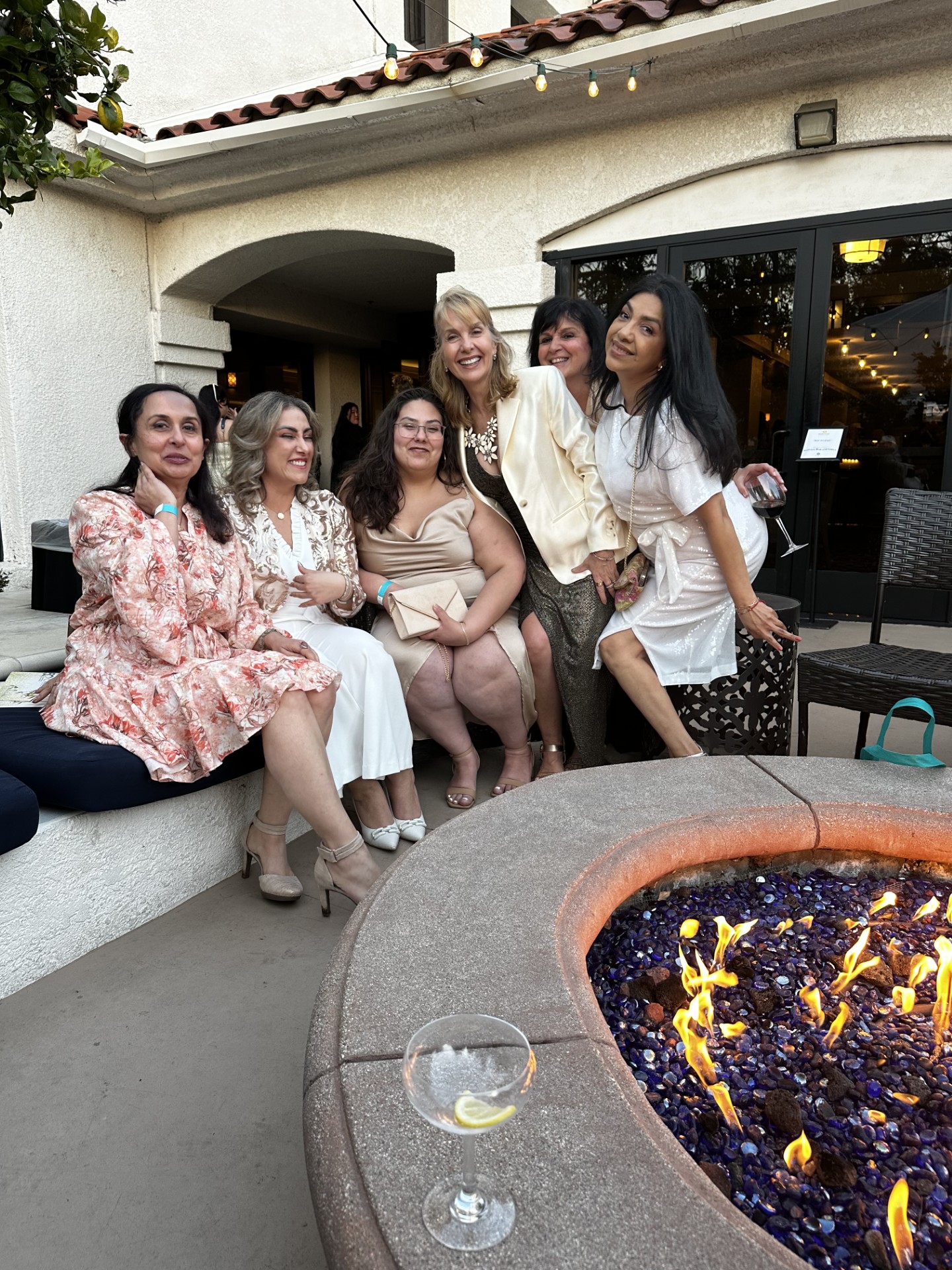 Group of six women sitting and standing outdoors near a fire pit, smiling, with a building in the background.