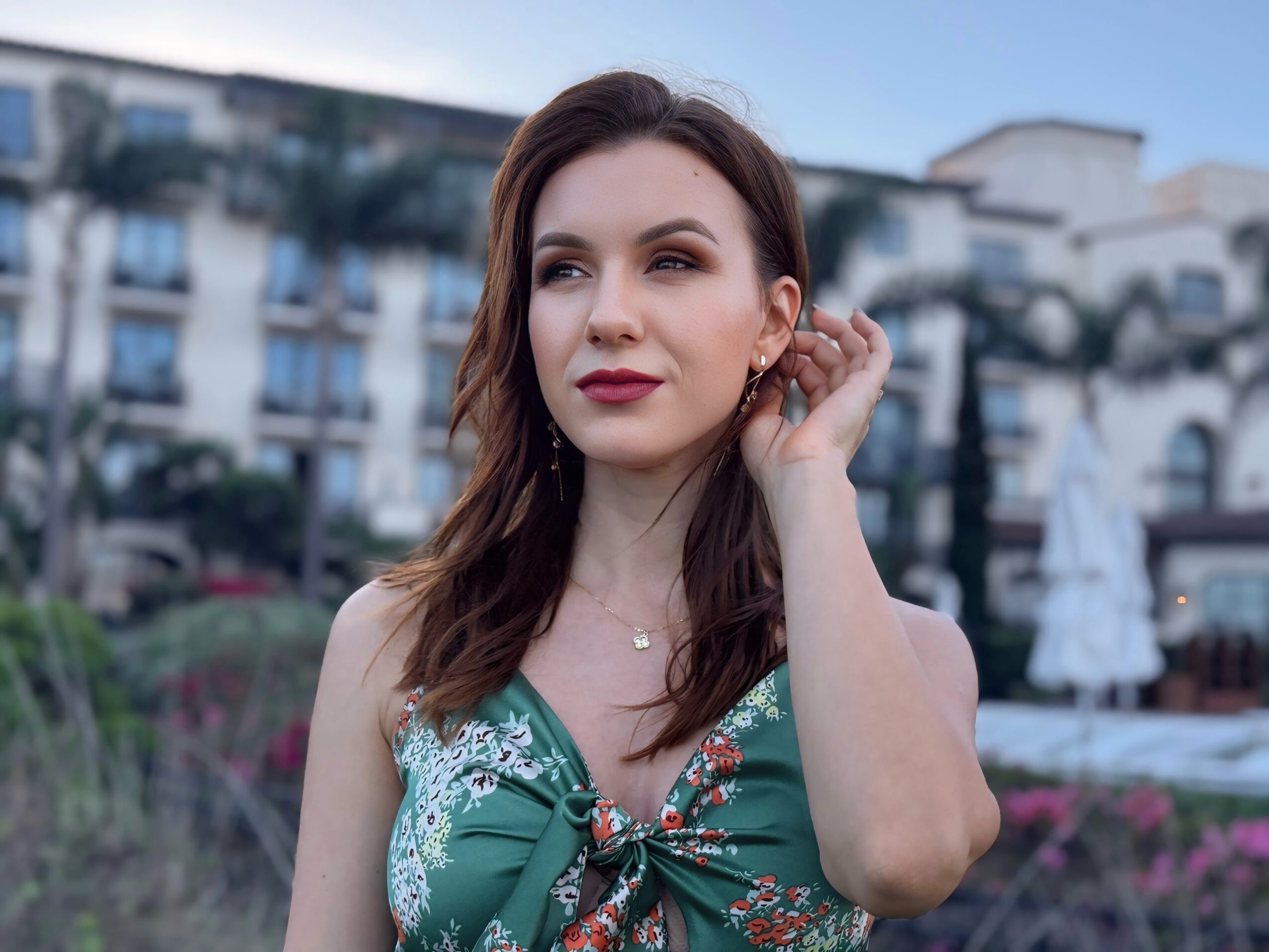 Young woman with brown hair and red lipstick, wearing a green dress outdoors, touching her ear, with buildings and flowers in background.