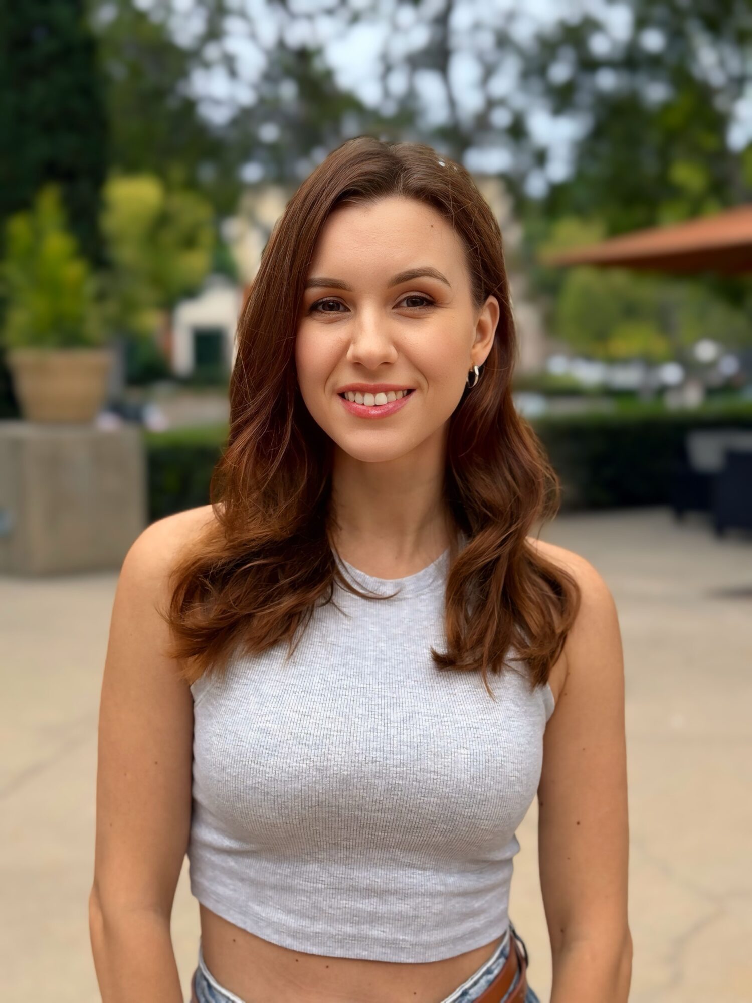 Young woman with long wavy brown hair smiling outdoors, wearing a sleeveless light gray top.