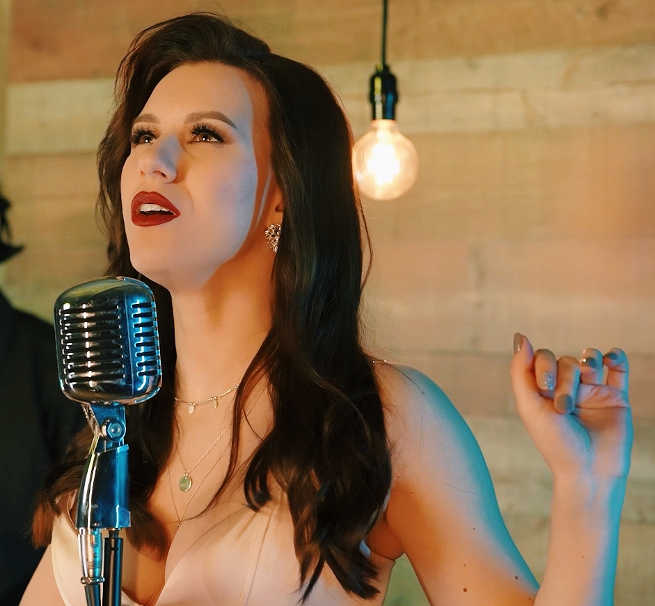 Woman with dark hair and red lipstick singing into a vintage microphone, gesturing with her right hand, in a warmly lit room.