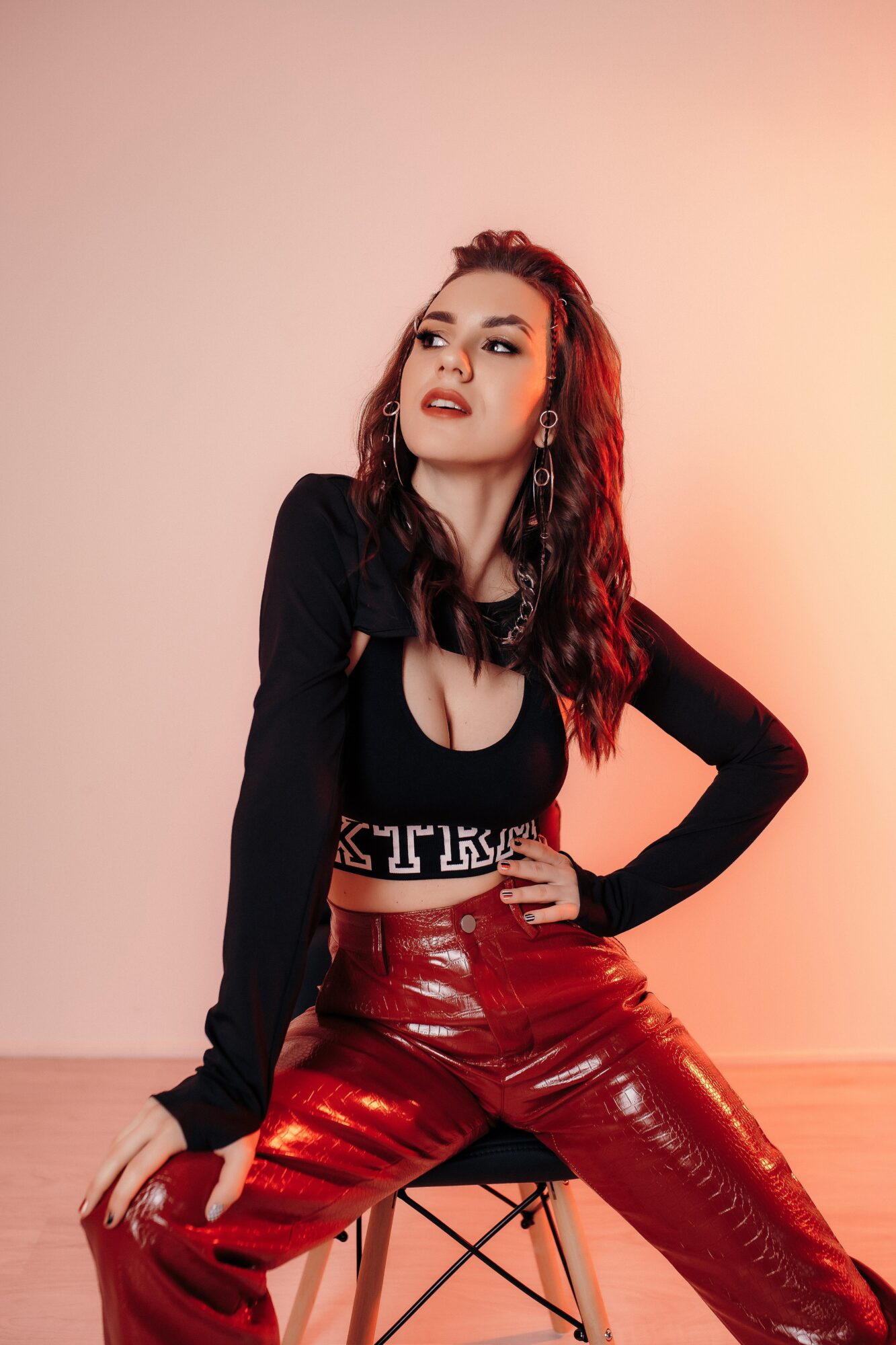 Woman with long wavy hair wearing a black top and red shiny pants, sitting on a stool against a plain background.