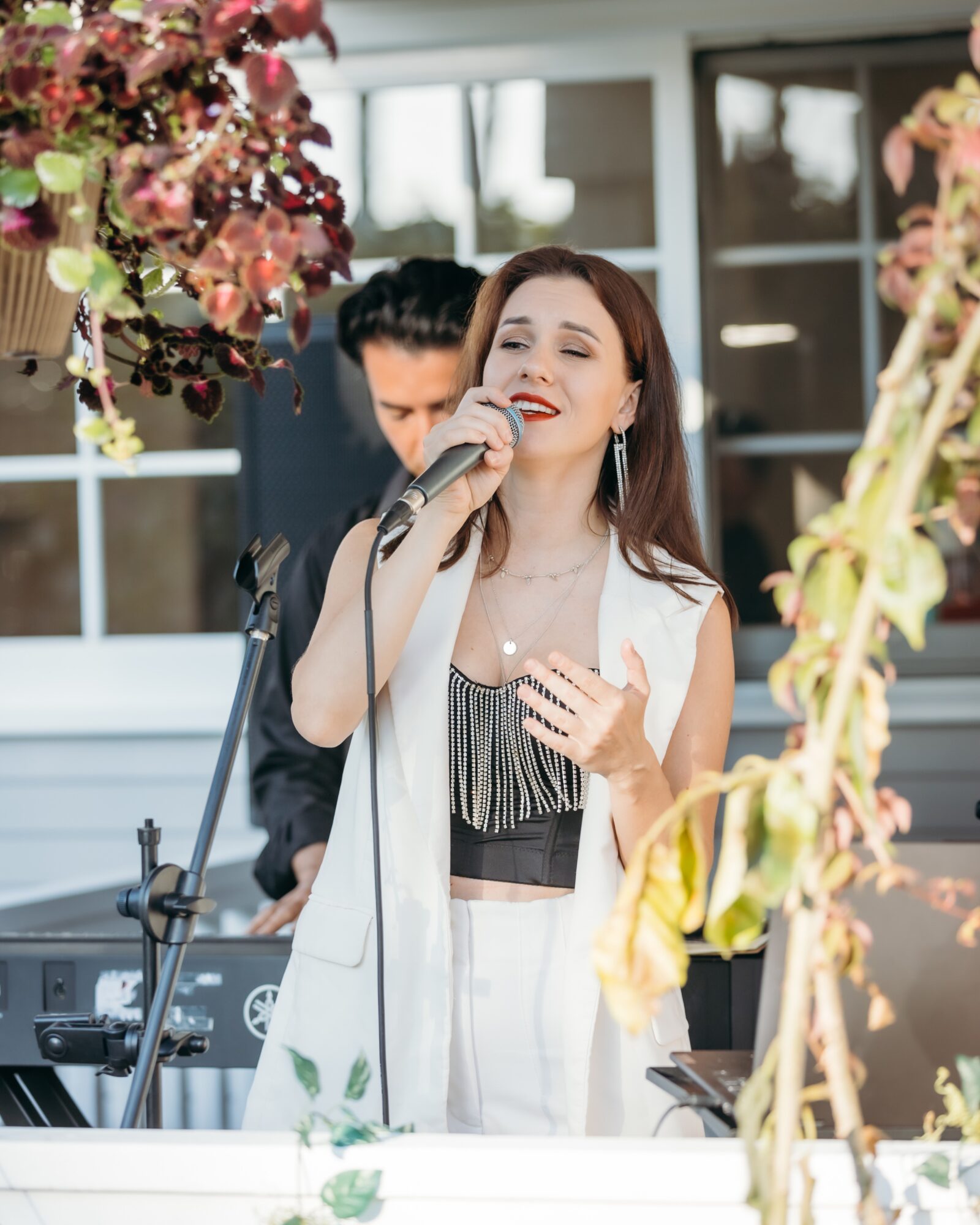 Woman singing into a microphone outdoors with a man playing keyboard behind her, surrounded by flowers.