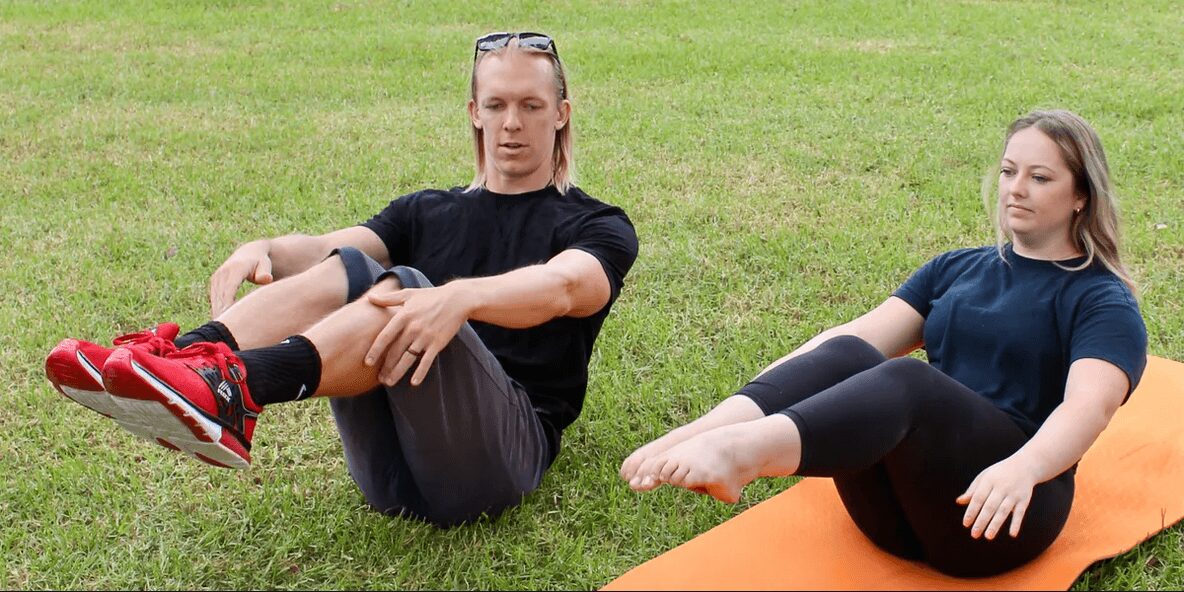 Two women sitting on grass, one on an orange mat, stretching legs, outdoors, daytime.