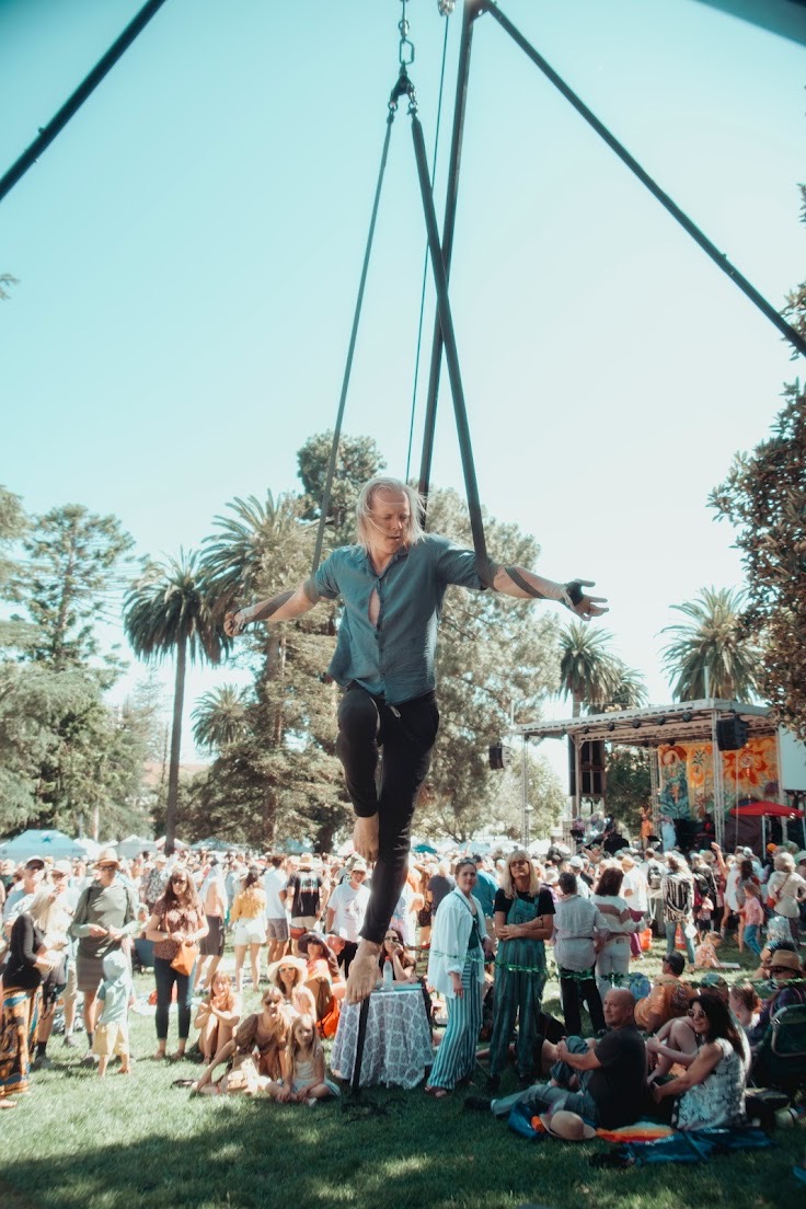 Person balancing on a large swing at an outdoor event with a crowd and palm trees in the background.