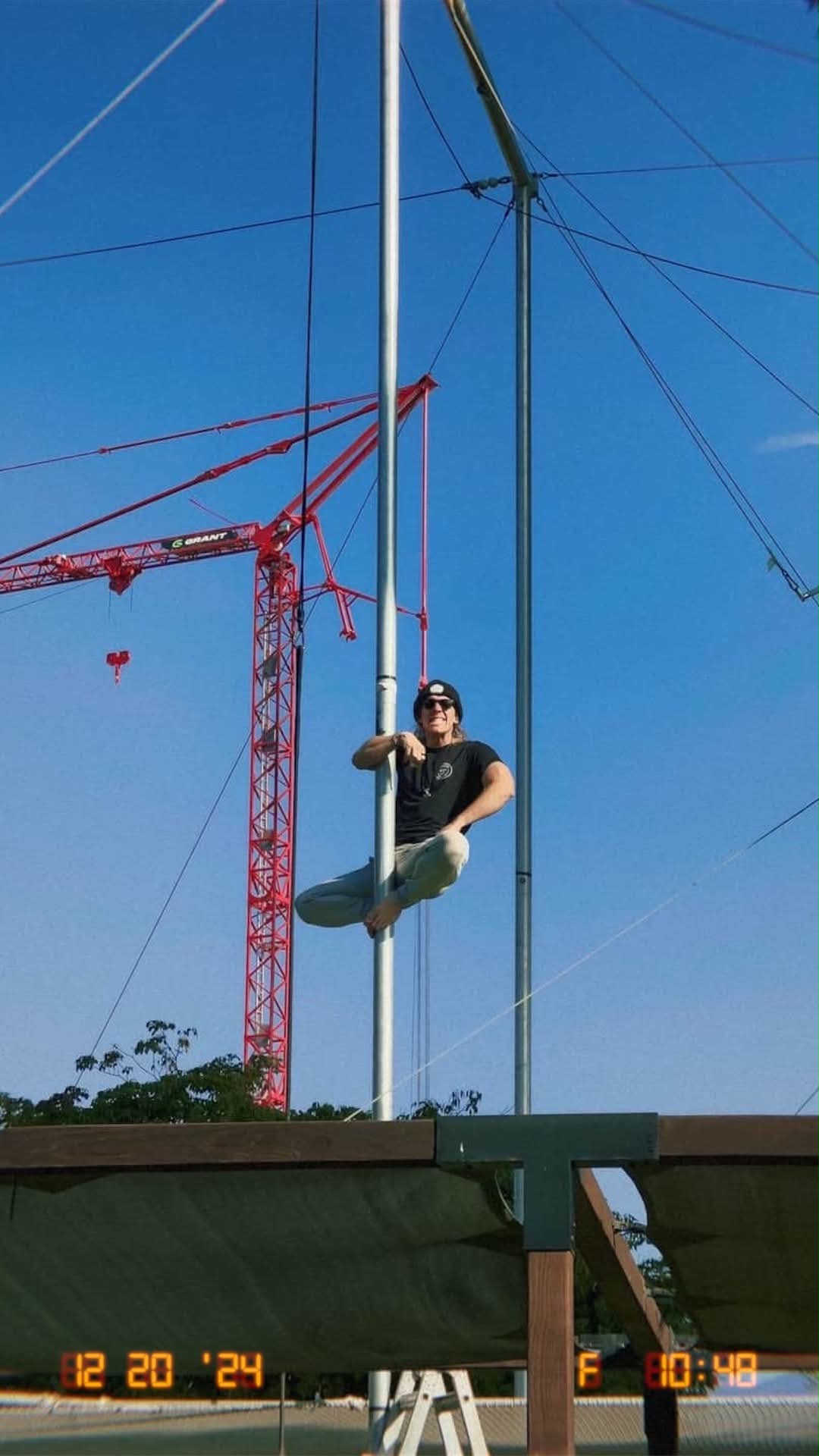 Person sitting on a swing hanging from a tall pole outdoors, with a construction crane in the background, blue sky.