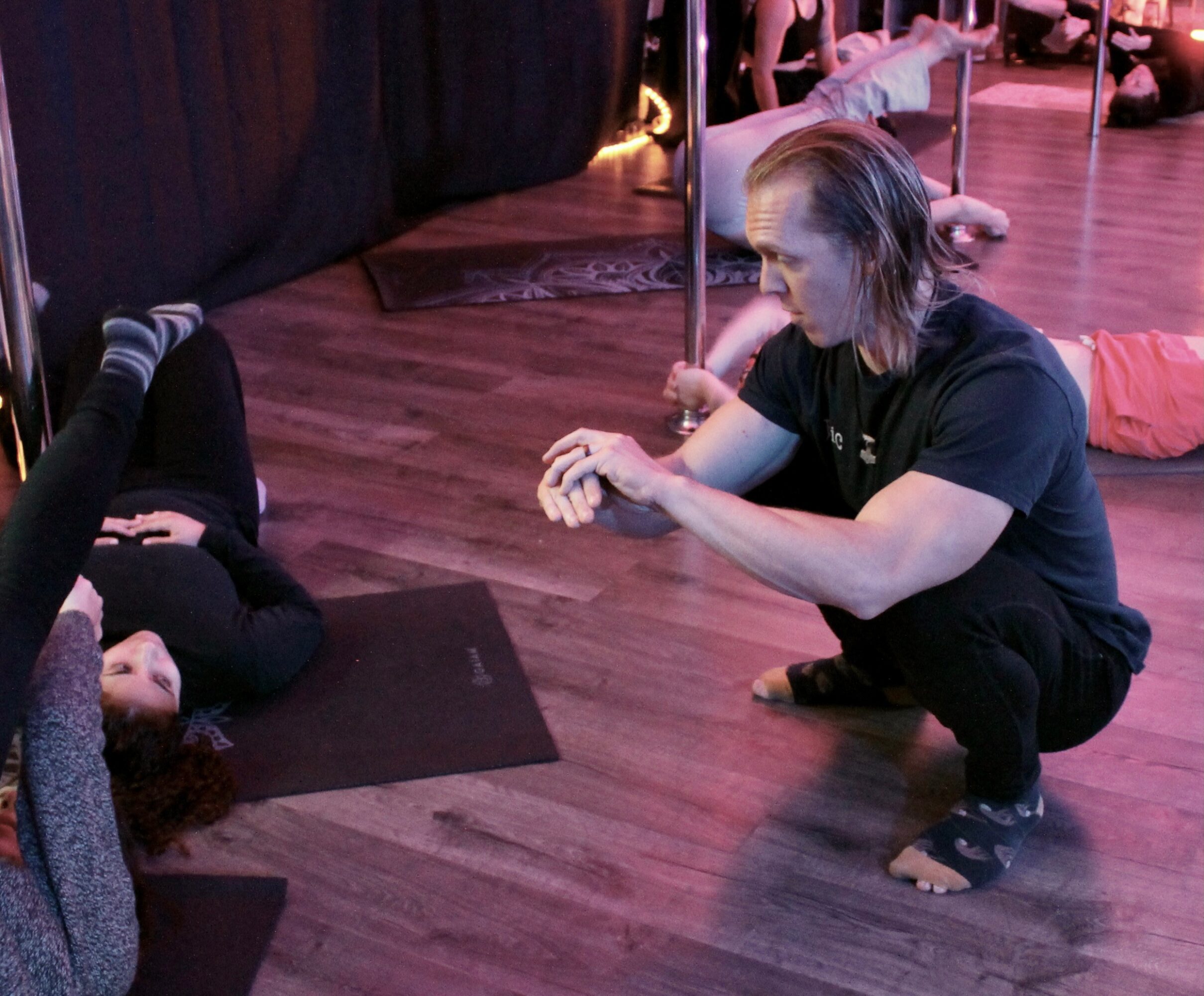 Person crouching on the floor with arms extended, others lying on mats nearby in a room with wooden floor and pink lighting.