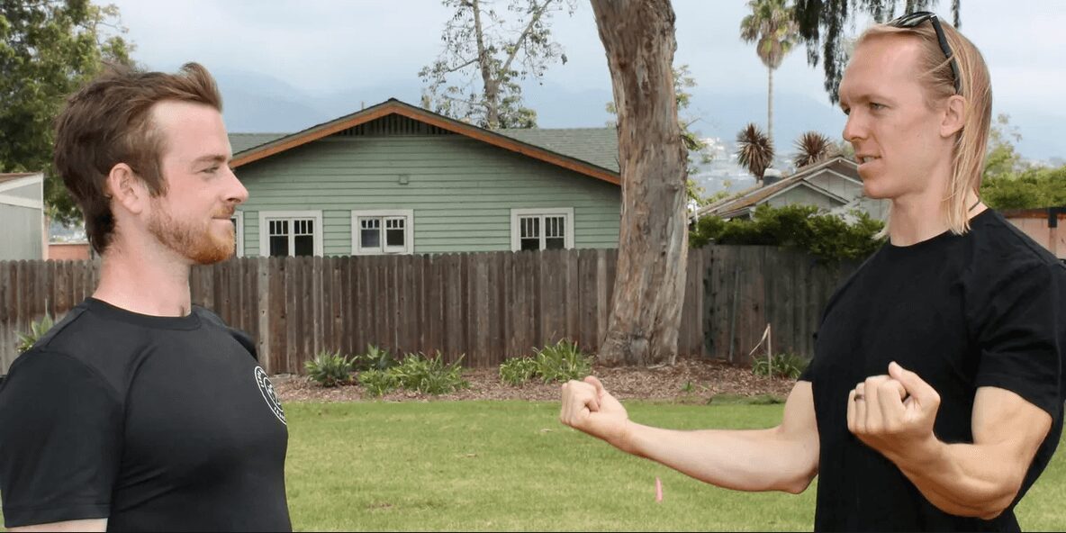 Two men face each other outdoors, one with a beard and the other with long hair, both in black shirts, fists raised.