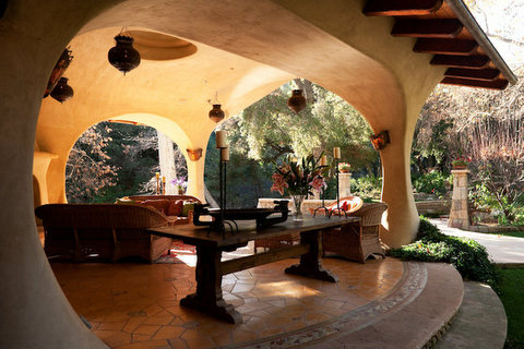 Open-air patio with curved arches, outdoor furniture, and lush greenery, sunlight filtering through trees, and a tiled floor.