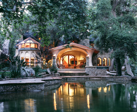 House with arched windows and a porch, surrounded by trees, reflected in a pond, evening lighting, lush greenery.