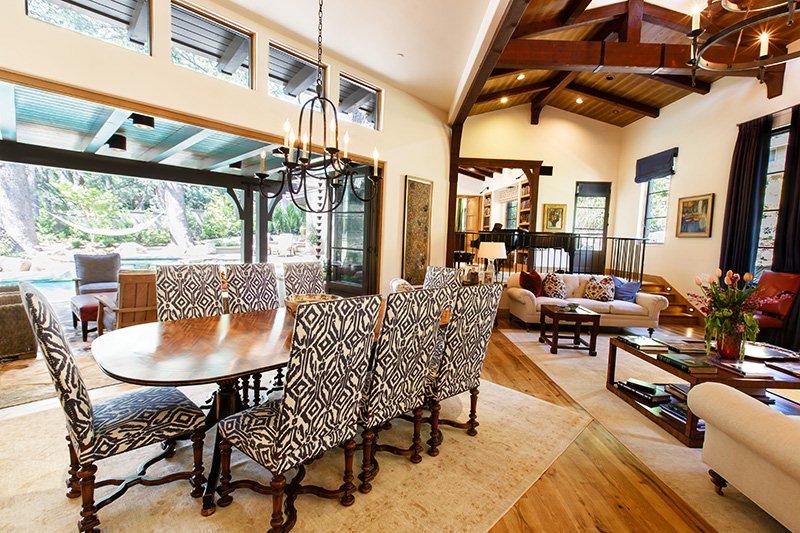 Living and dining area with large window, wooden furniture, patterned chairs, and a high ceiling with exposed beams.