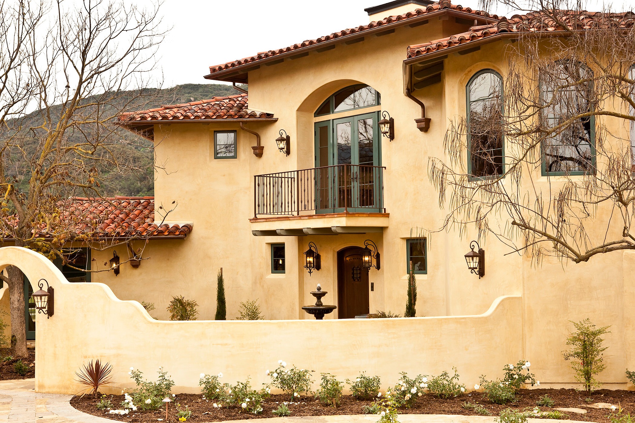 Two-story house with a beige stucco exterior, red tile roof, arched windows, and a small balcony, surrounded by a garden.