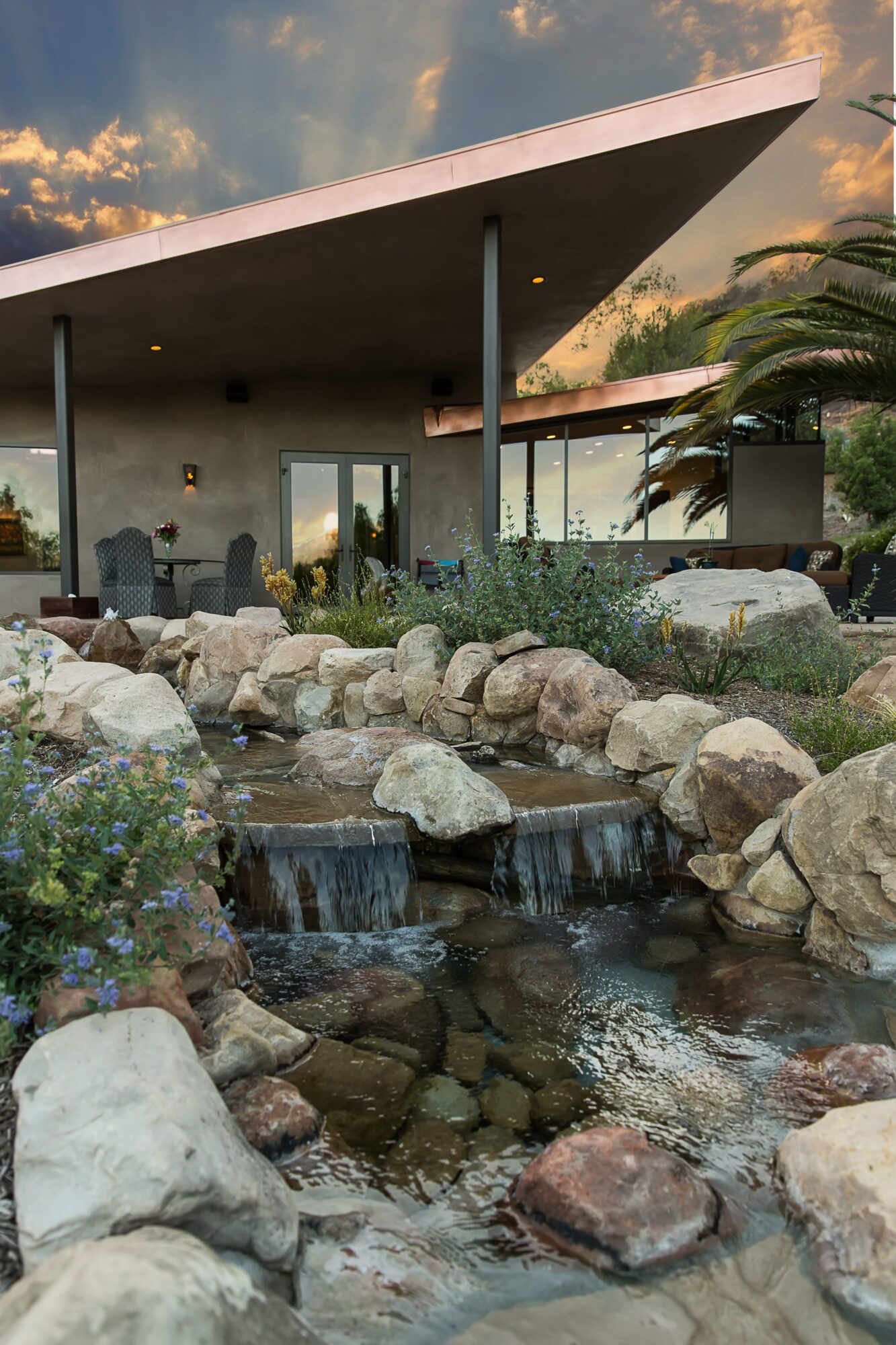 Modern house with large overhanging roof, surrounded by rocks, plants, and a small waterfall in the foreground.